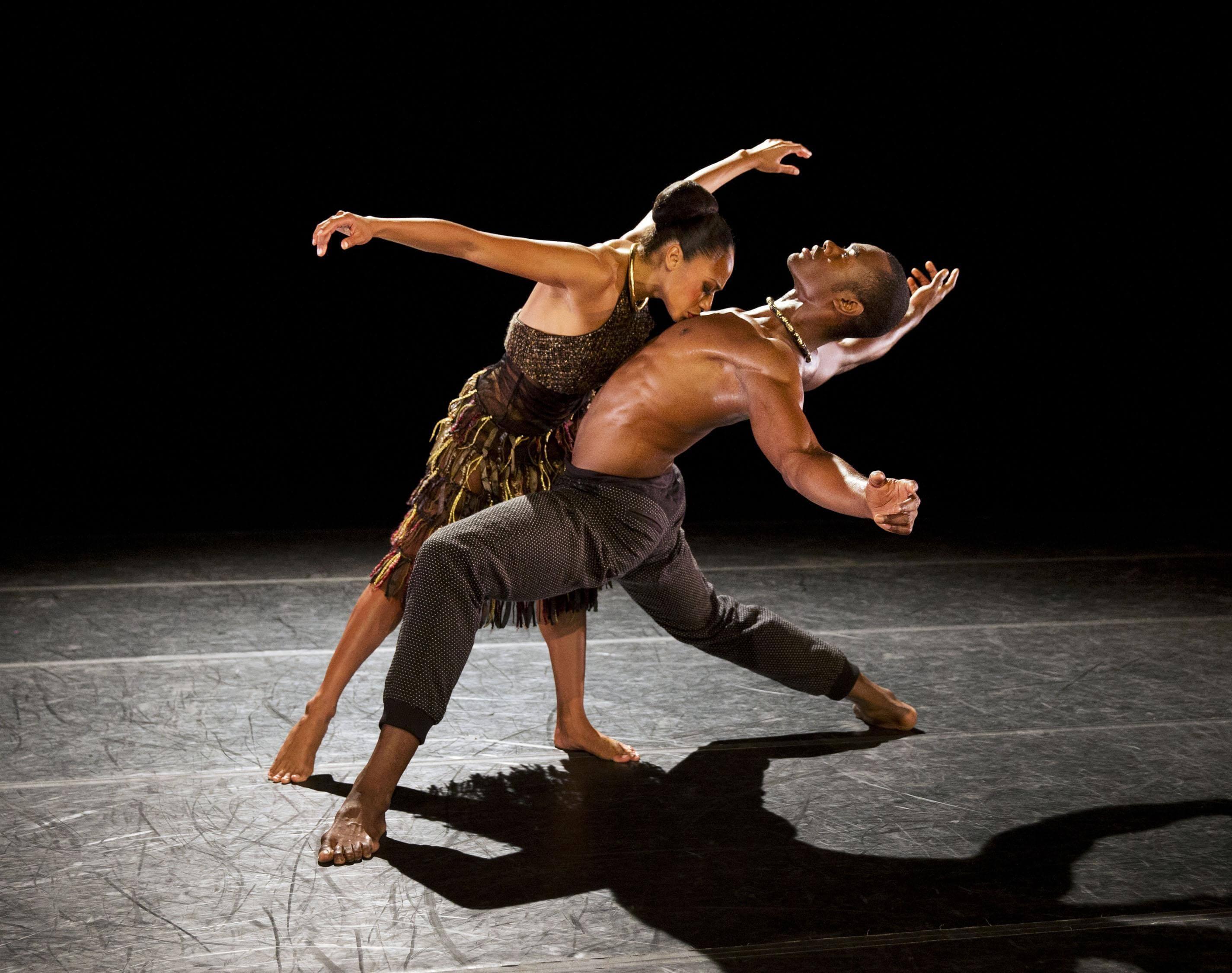 A male and female dancer on stage, the male dancer leaning back with one leg bent, while the female dancer arches over him with arms extended, both against a dark background.