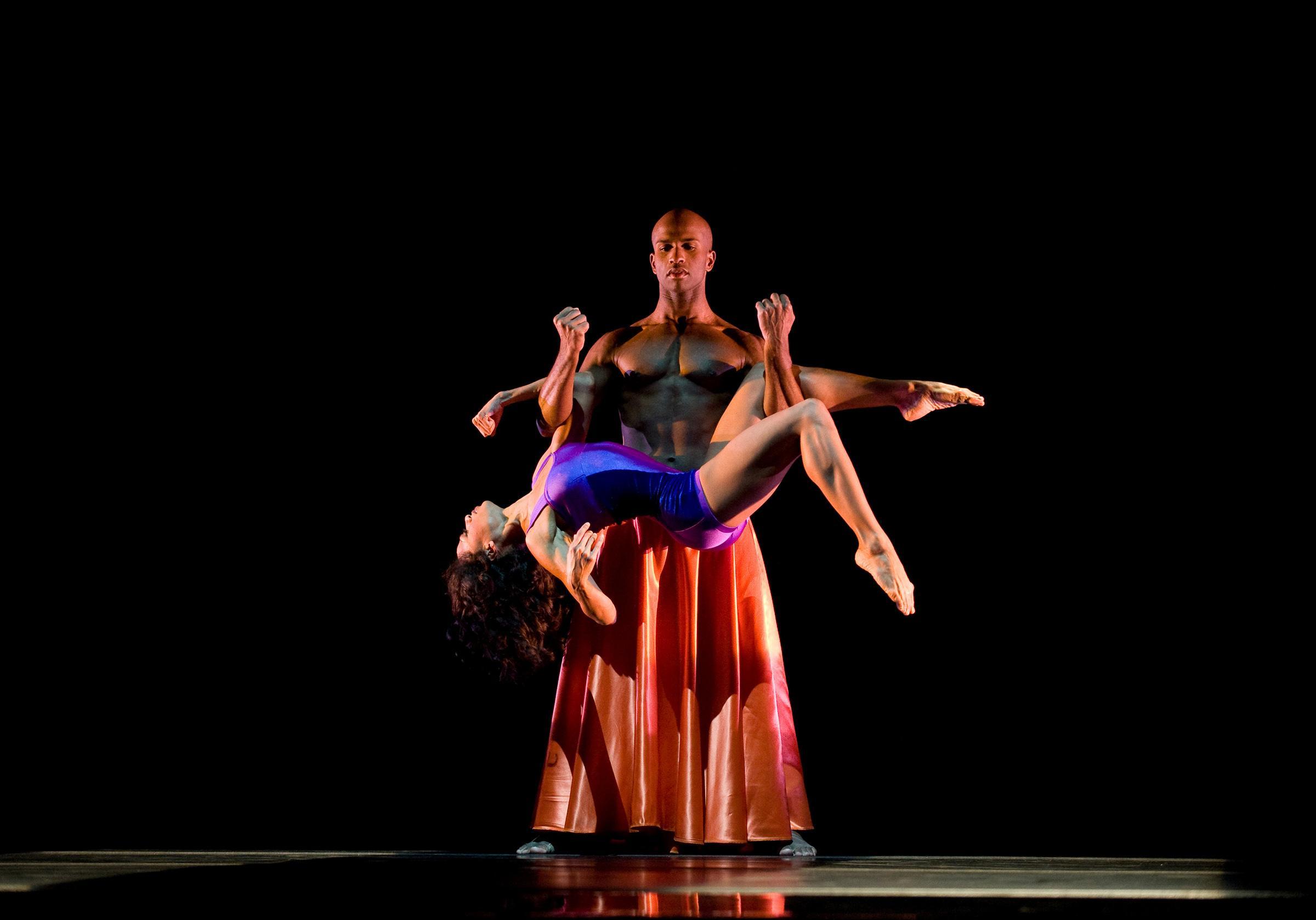 A male dancer wearing a long orange skirt holds a female dancer in a purple leotard above his head, her body arched backward. The background is black.