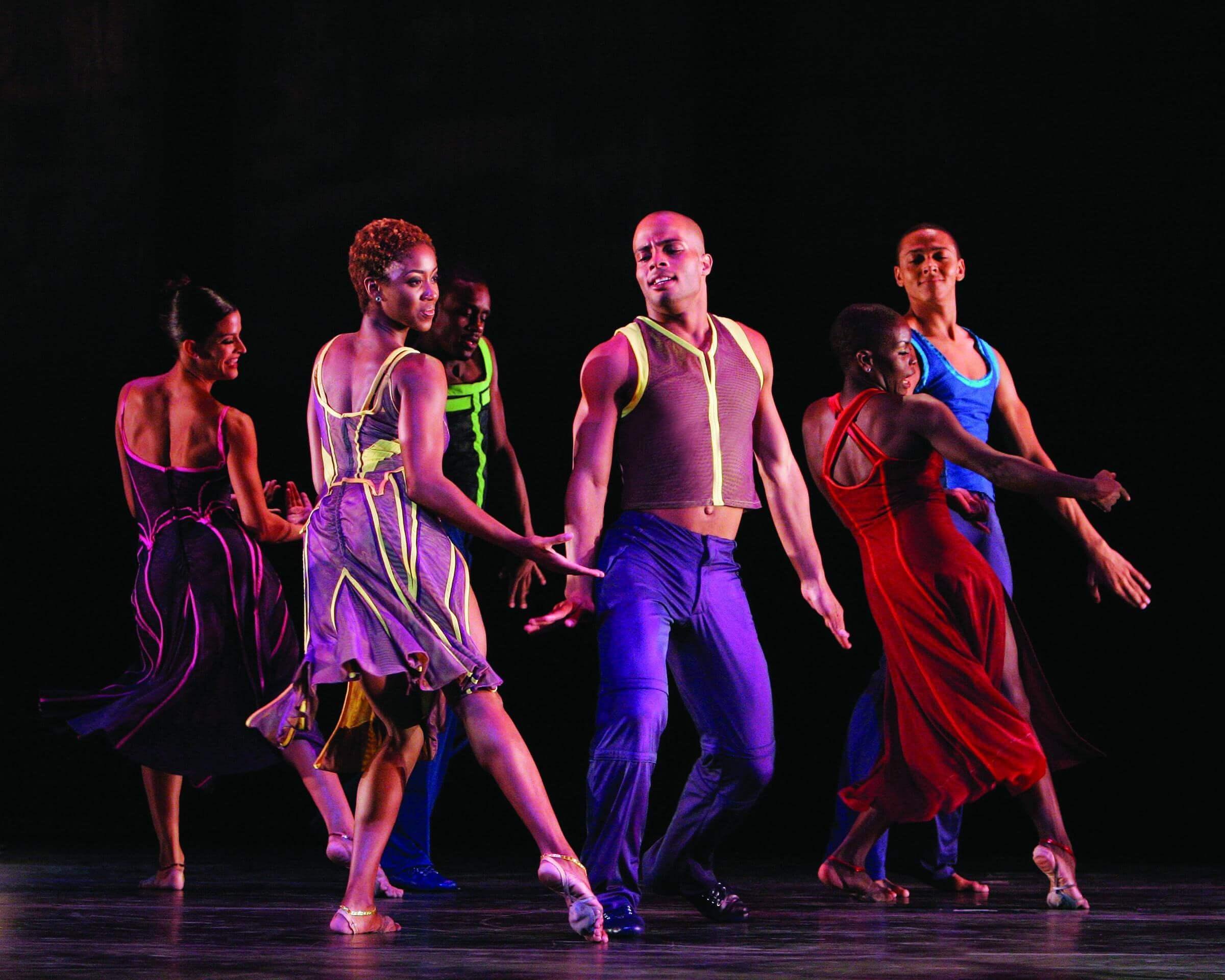  Six dancers in colorful costumes perform on stage against a black background. The dancers are in motion, with some turning and others extending their arms, showcasing their fluid movements and vibrant attire.