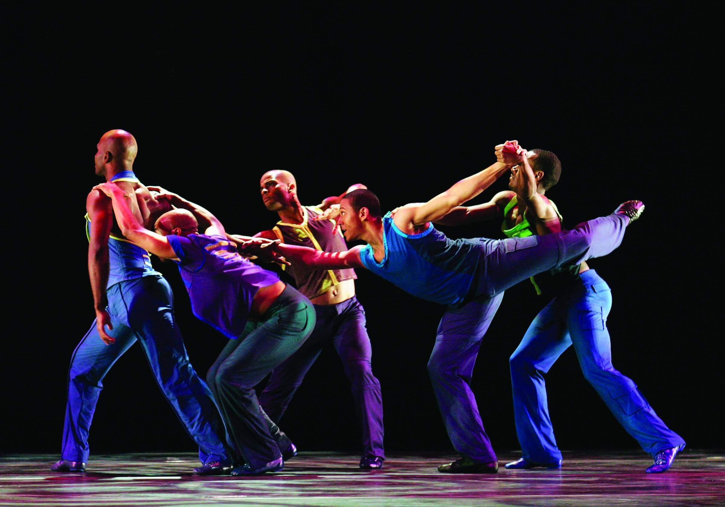 Six dancers in brightly colored outfits perform a dynamic routine on stage against a black background. The dancers are engaged in a complex, intertwined formation, showcasing their strength and flexibility.