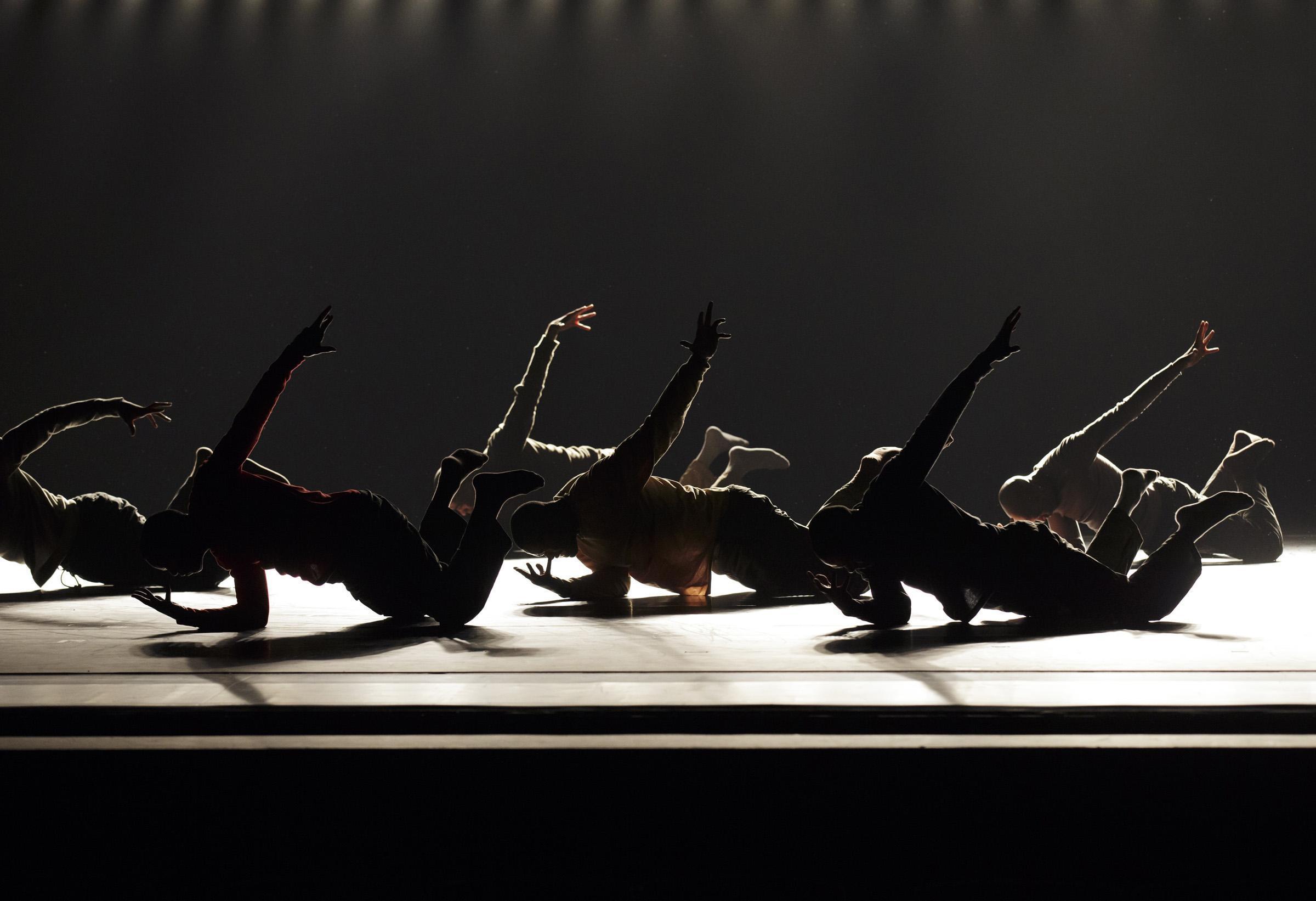 Six dancers on stage, lying on their sides with one arm extended upward and the other supporting them on the ground. The dancer in the center wears a red shirt, while the others wear shades of beige and blue. Bright stage lights form a row in the background.