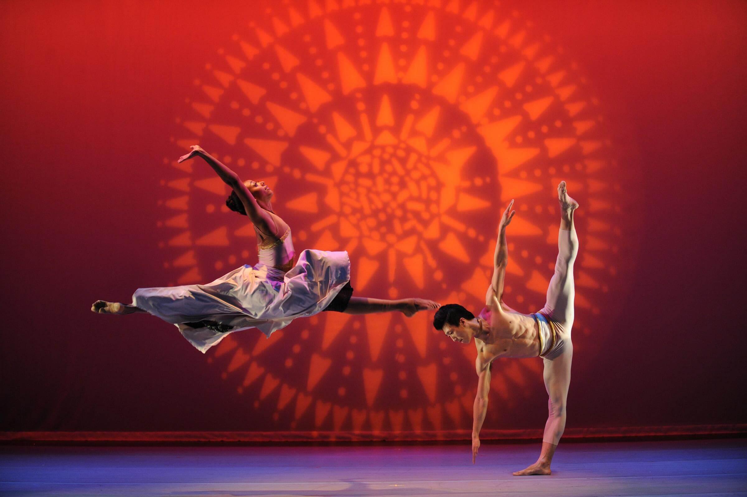 Two dancers, one leaping in the air and doing a split, the other perpendicular to the ground, with both arms and legs straight and extended apart from one another