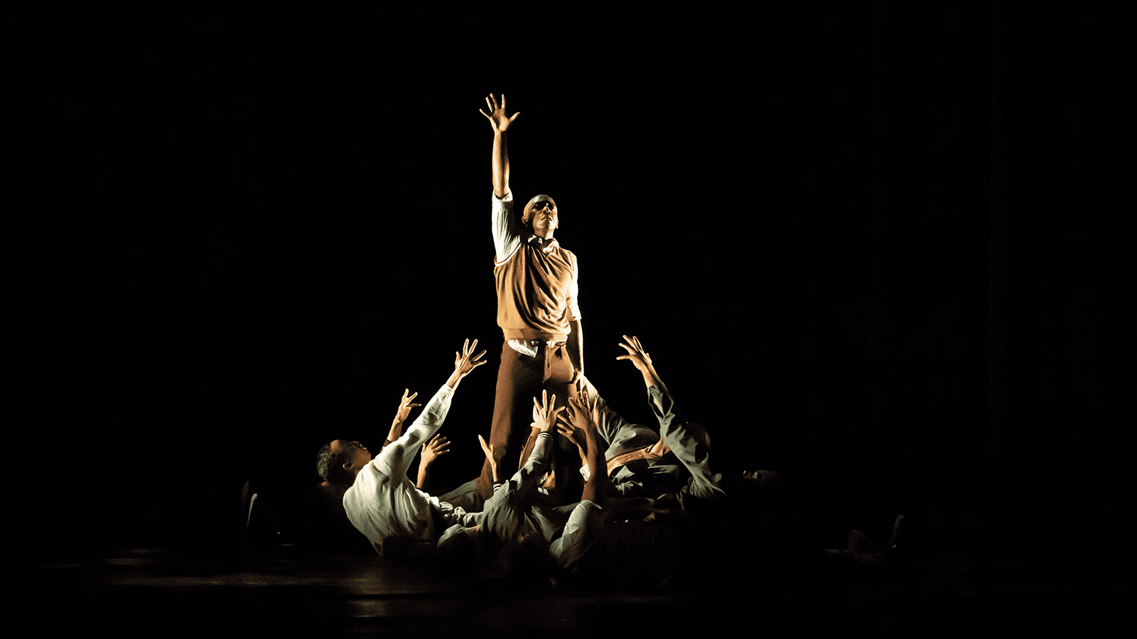 A dancer reaching one of his arms into the air. The dancers lower than him are raising their arms towards him