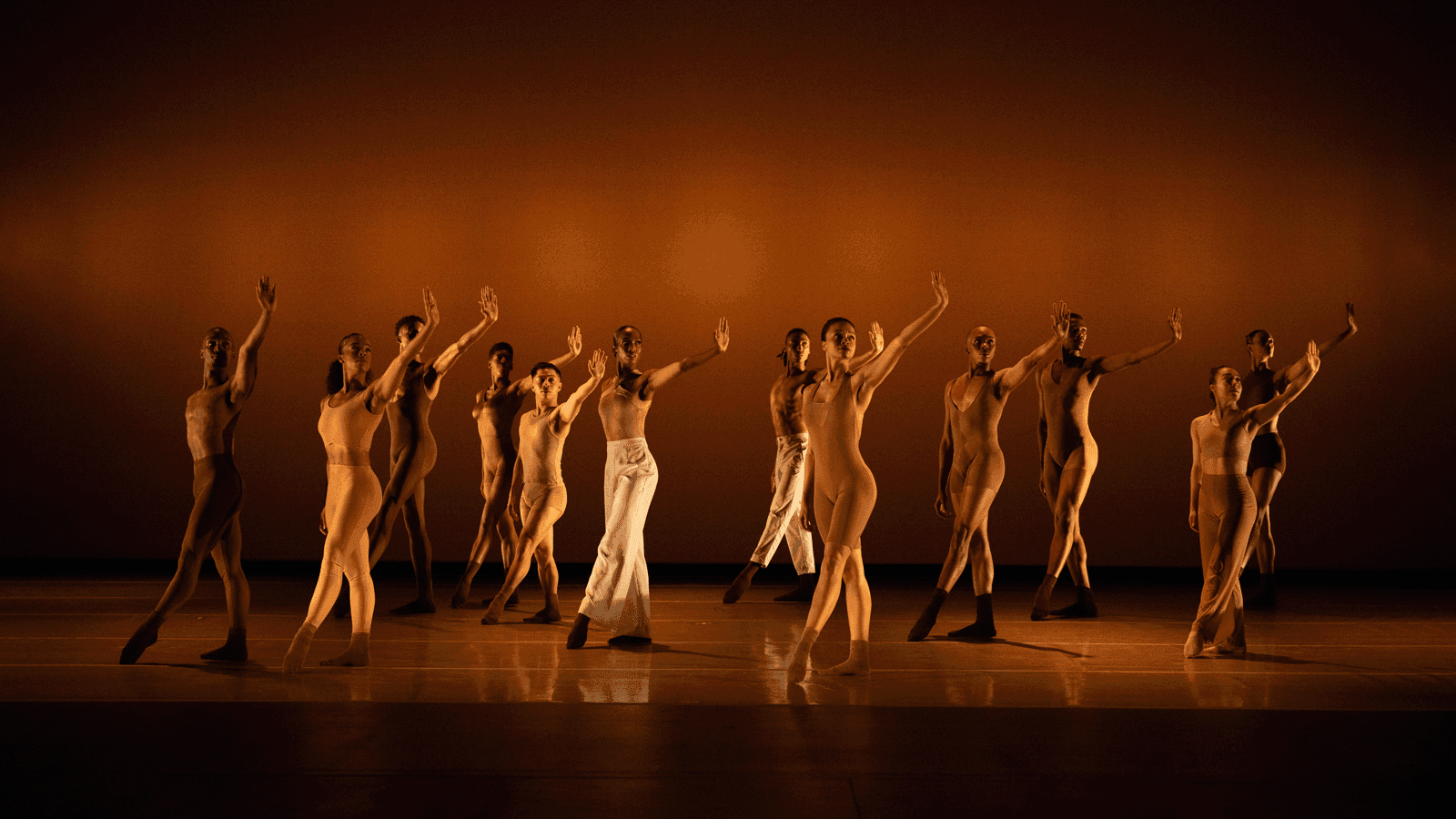 Group of dancers in beige and brown costumes standing in a line, each with one arm raised, against an orange background.