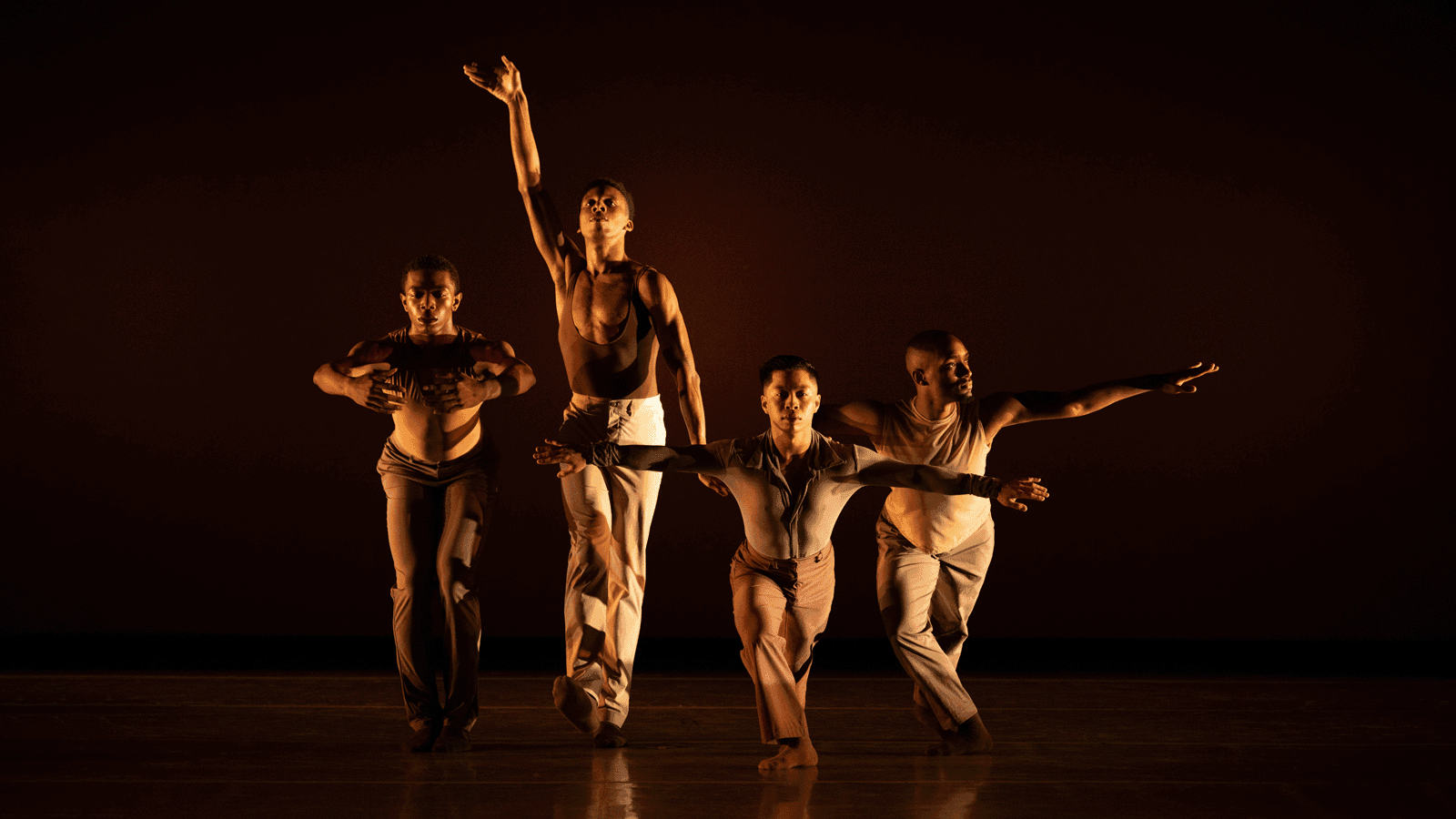 Four dancers in various poses on stage, wearing beige and brown costumes, against a dark background. One dancer has an arm raised high, another extends arms forward.