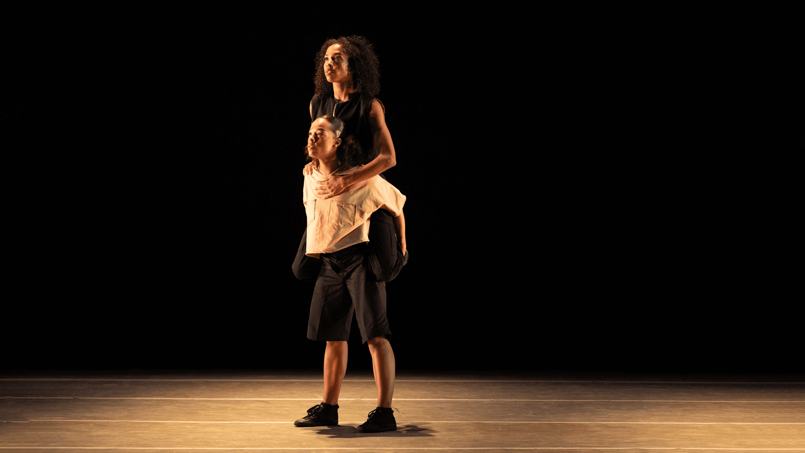 One dancer stands while supporting the other dancer seated on their shoulders, both wearing loose-fitting outfits. They perform on a dimly lit stage, creating a dramatic and focused visual.