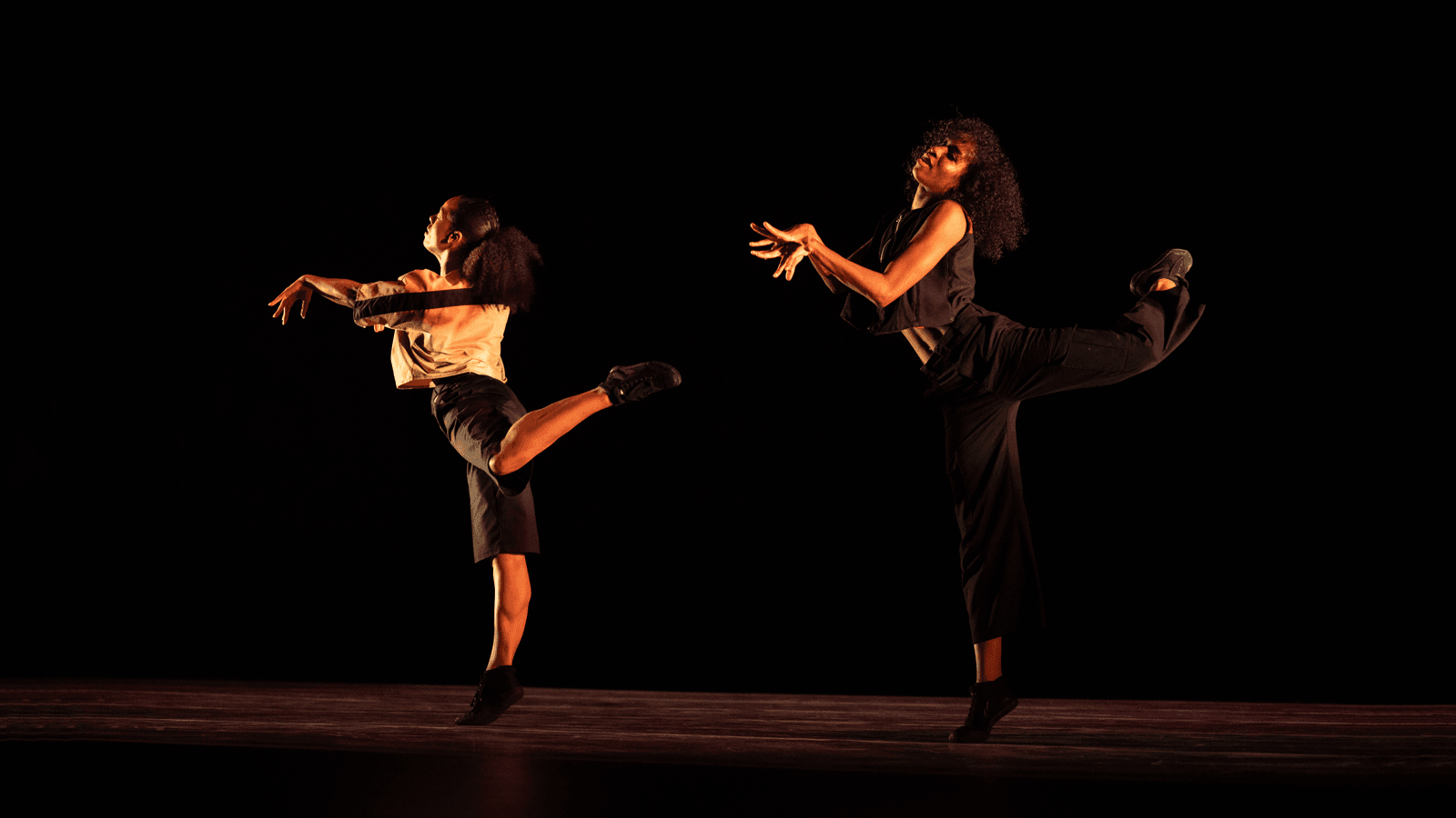 Two dancers perform on a dark stage, each balancing on one leg with the other leg extended behind them. They wear loose-fitting outfits, and their arms are gracefully positioned, adding to the expressive quality of the performance.