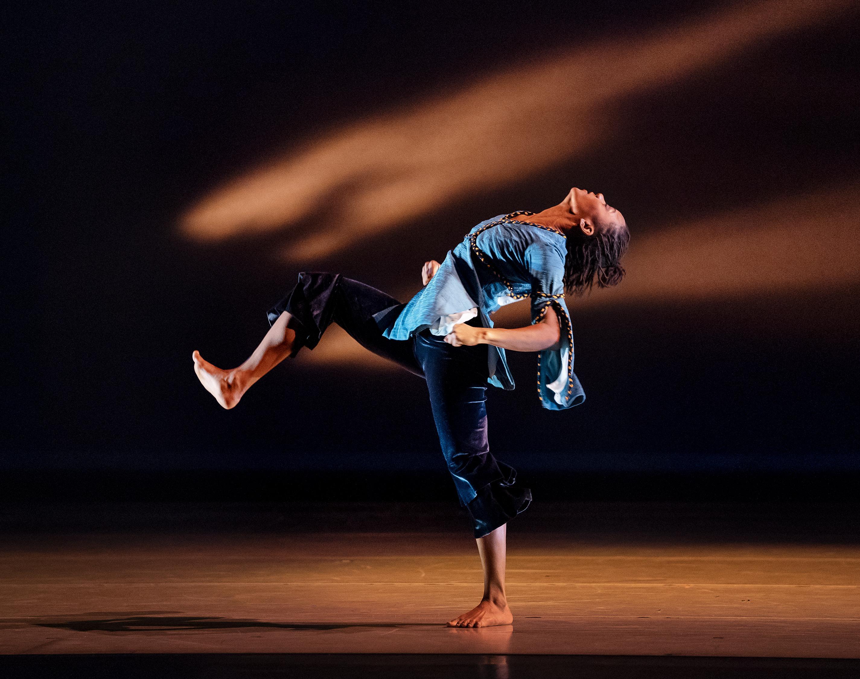 A woman is dancing on stage. Her head is tilted back and one knee is in the air.