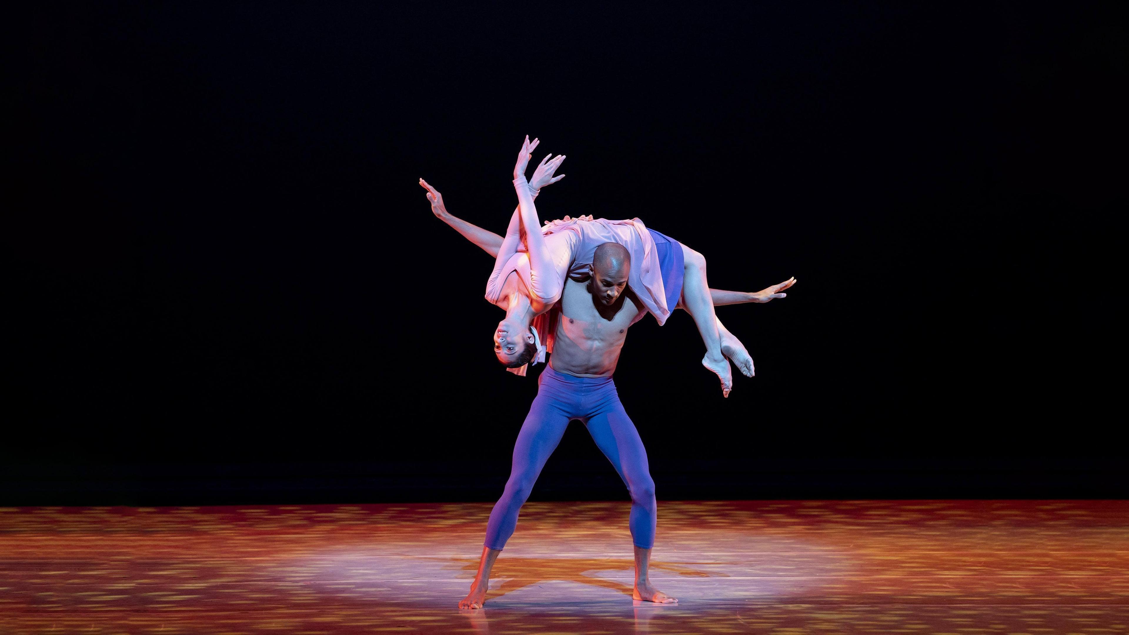 Two dancers on stage. The man is standing with his arms outstretched and the woman is draped over his shoulders.