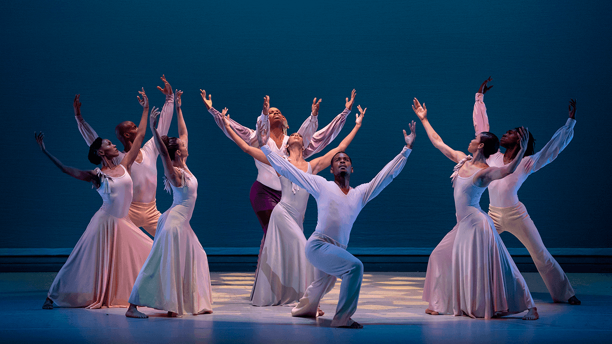 A group of dancers are on a blue stage posing with their arms outstretched in different directions