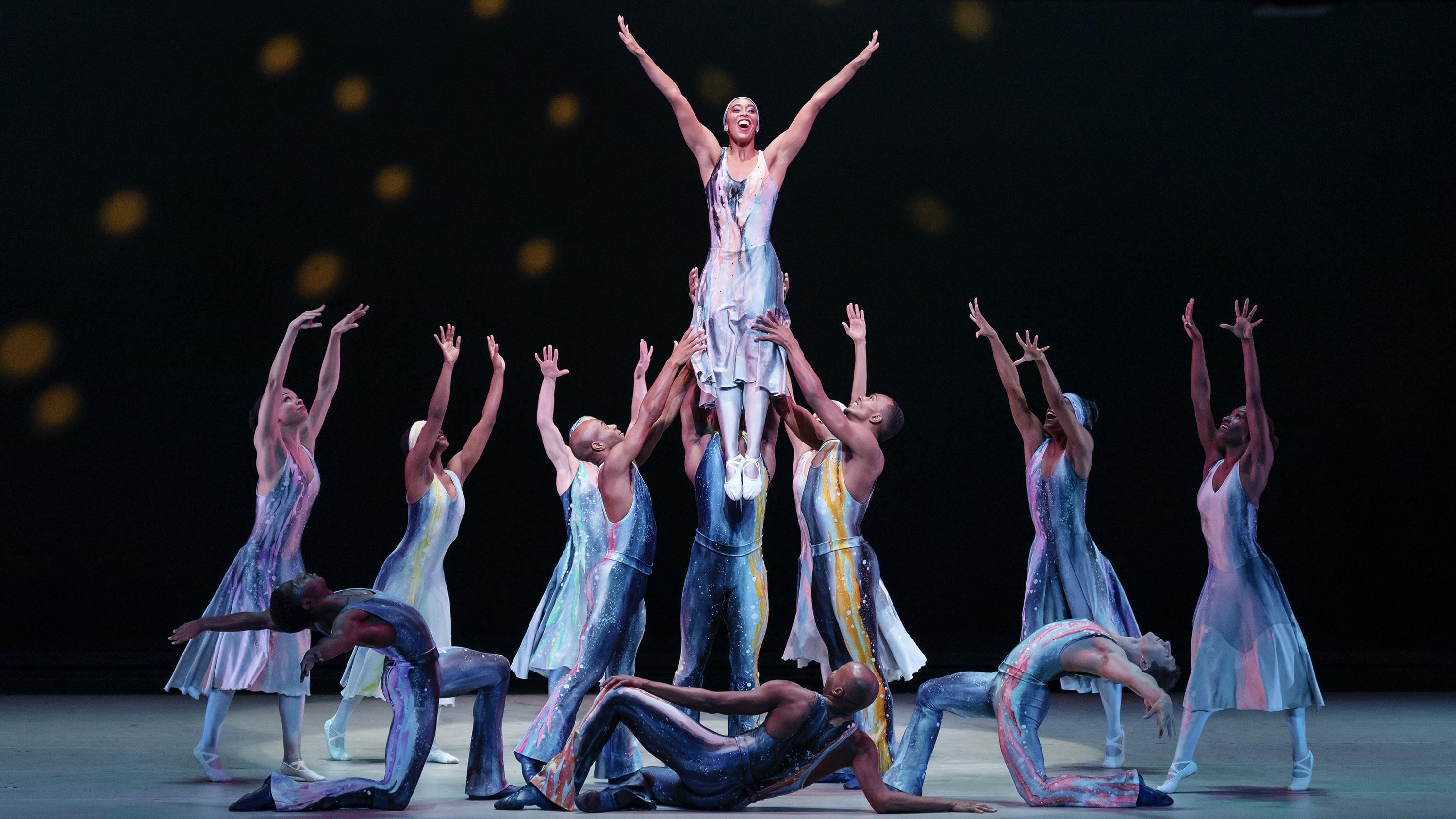 A group of dancers on stage in blue gradient costumes stand facing a dancer being lifted in the air. Everyone's arms are raised.