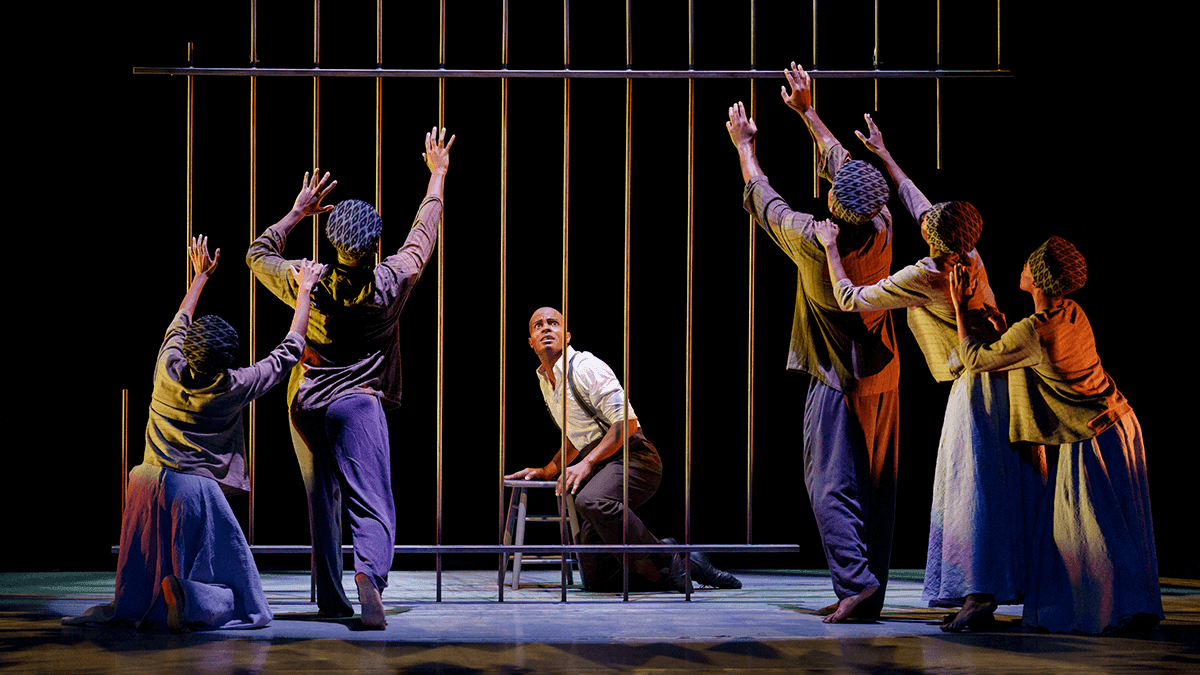 A color photo of the Alvin Ailey American Dance Theater performing Alvin Ailey's Survivors, captured by Paul Kolnik. The image features a group of dancers reaching towards vertical bars, with a central male dancer seated on a stool, looking up with an expression of hope or determination. The dancers surrounding him are in long skirts and head wraps, extending their arms upwards, creating a visually striking and powerful composition. The stage lighting enhances the dramatic effect, highlighting the emotional