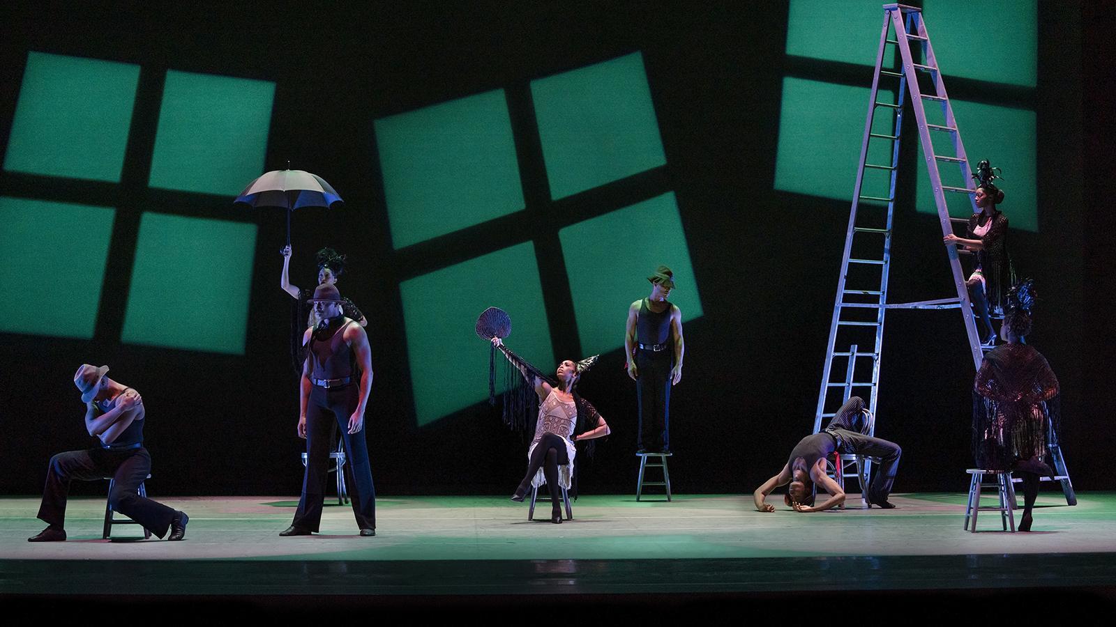 A group of dancers are on stage. There are green projected windows on the back and a ladder on the right. The dancers are strewn about in poses.