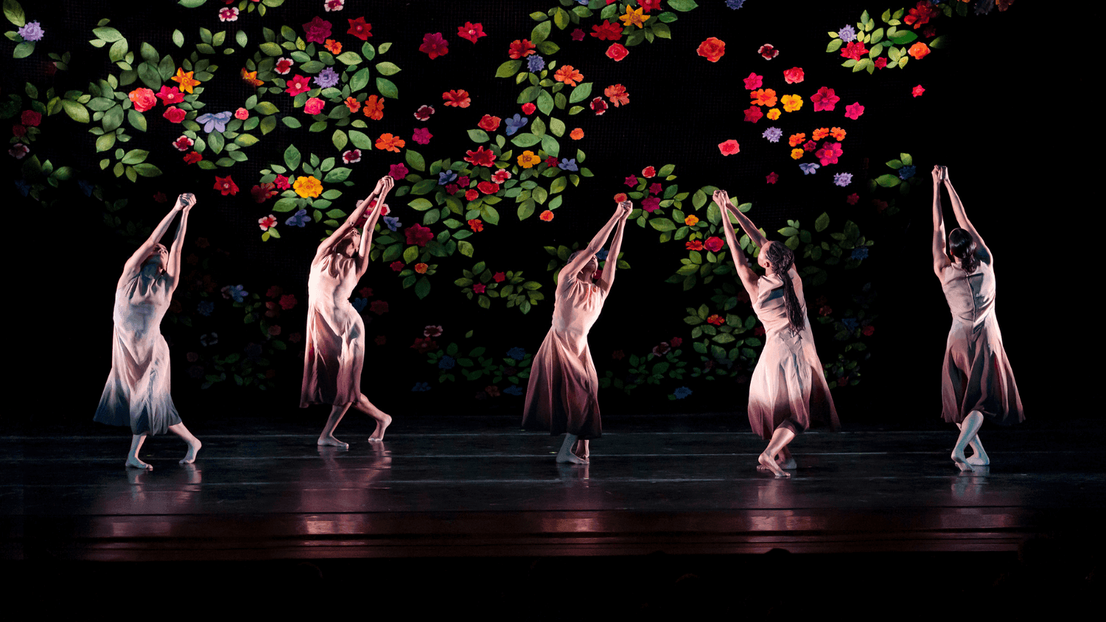 The ailey company all posing with their legs crossed and their arms reaching above their heads and touching. There is a floral pattern in the background.