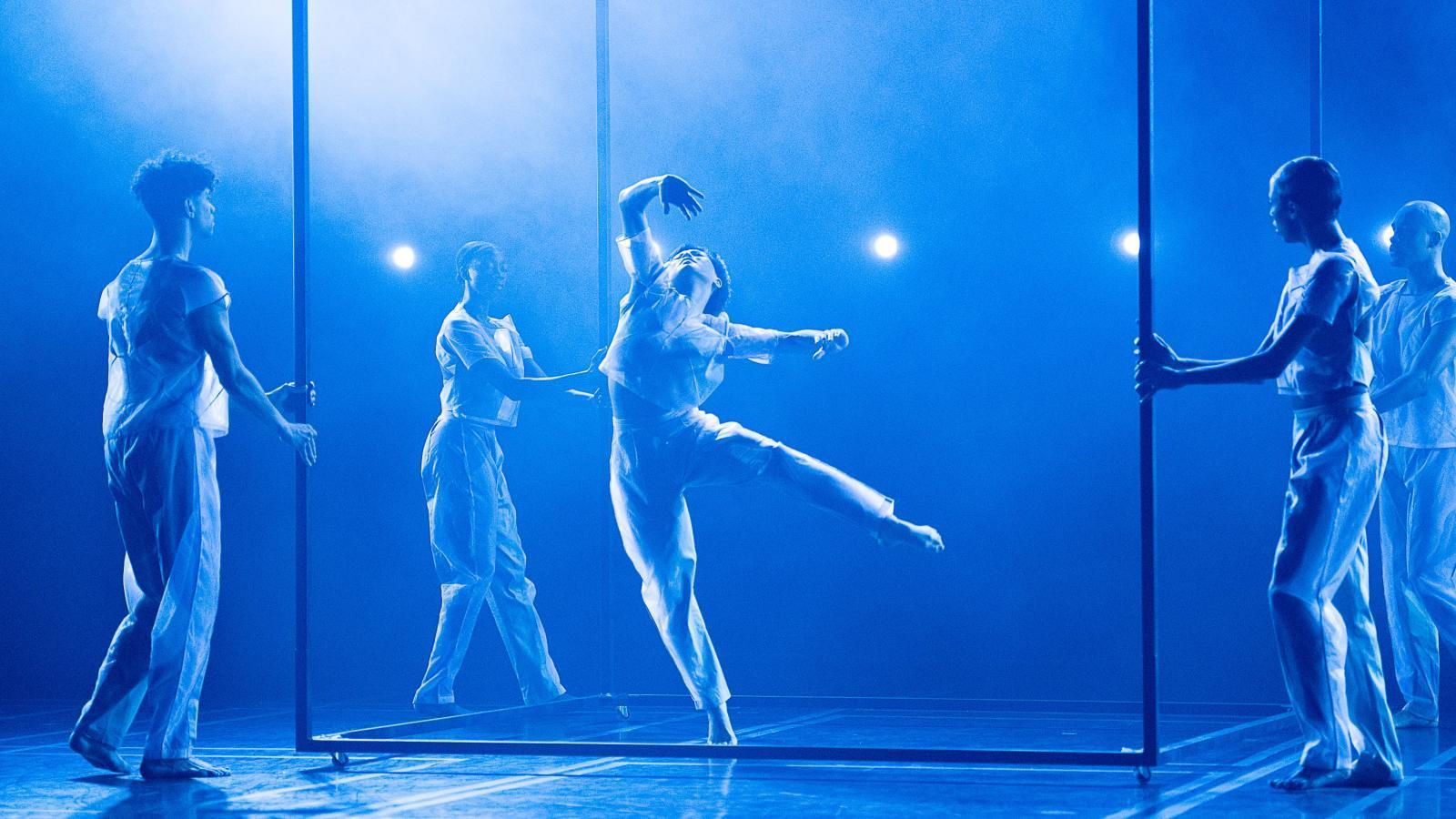 A bright blue light shines on the stage as four members of the company holds the ends of a transparent cube, while one member poses in the center