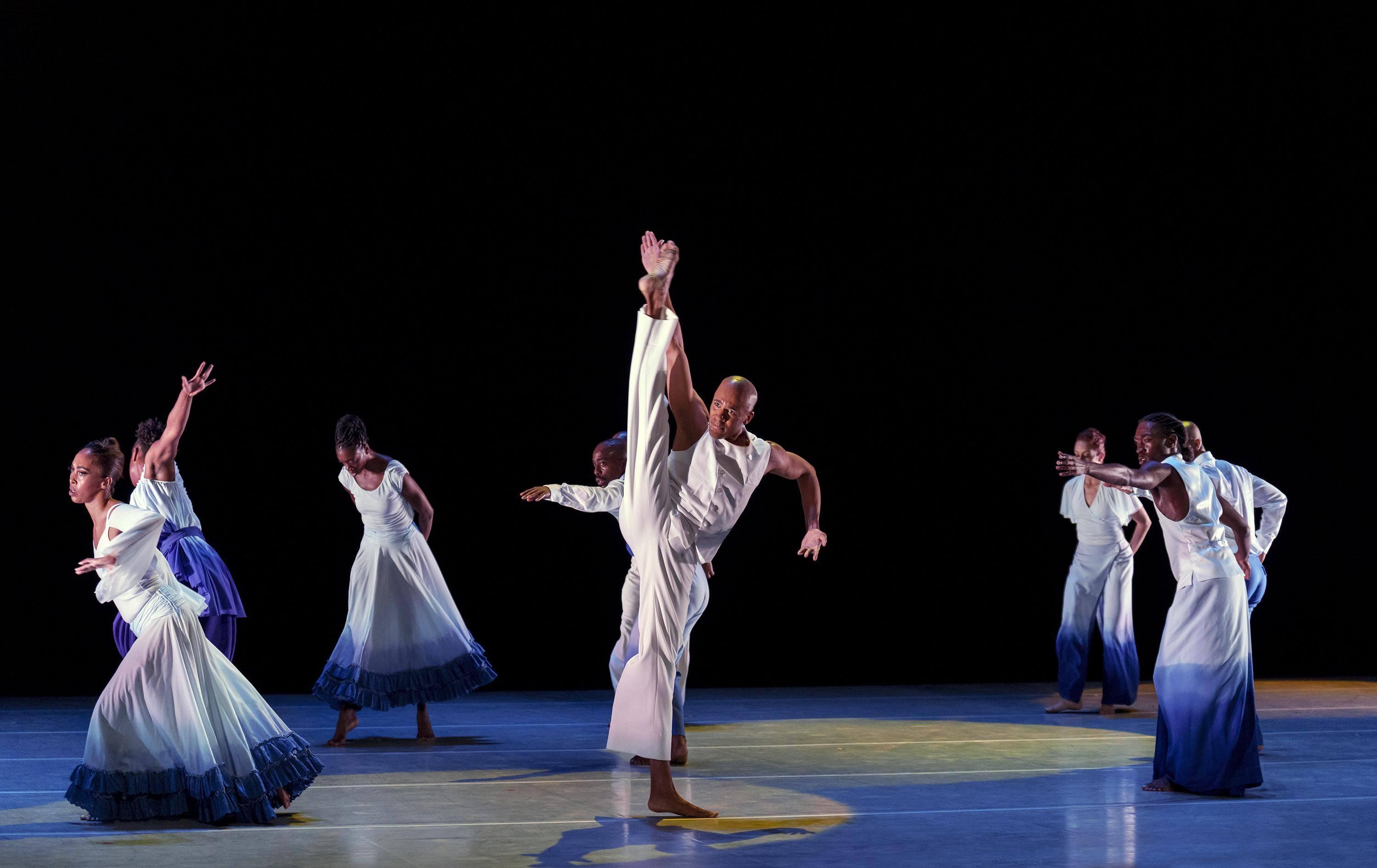 A dancer center stage raises one of his legs in the air performing a horizontal split. Members of the company surround this performer in a large circle