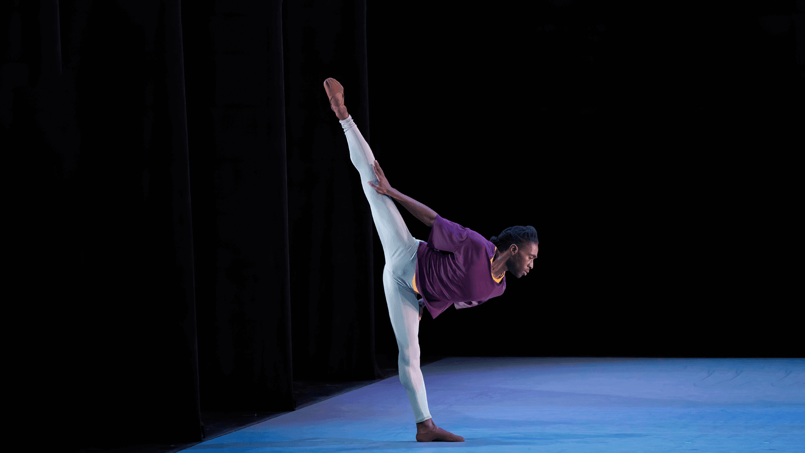 A man in a purple shirt and white pants has one leg in the air in an arabesque