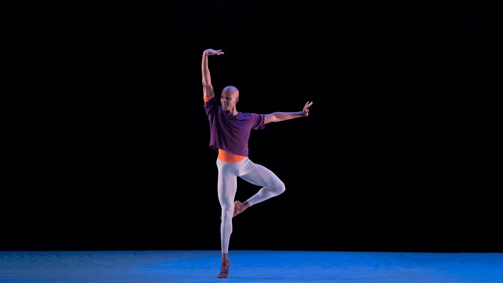 A man in a purple shirt and white pants on stage is on one leg with the other bent on his knee. His arms make an L shape.