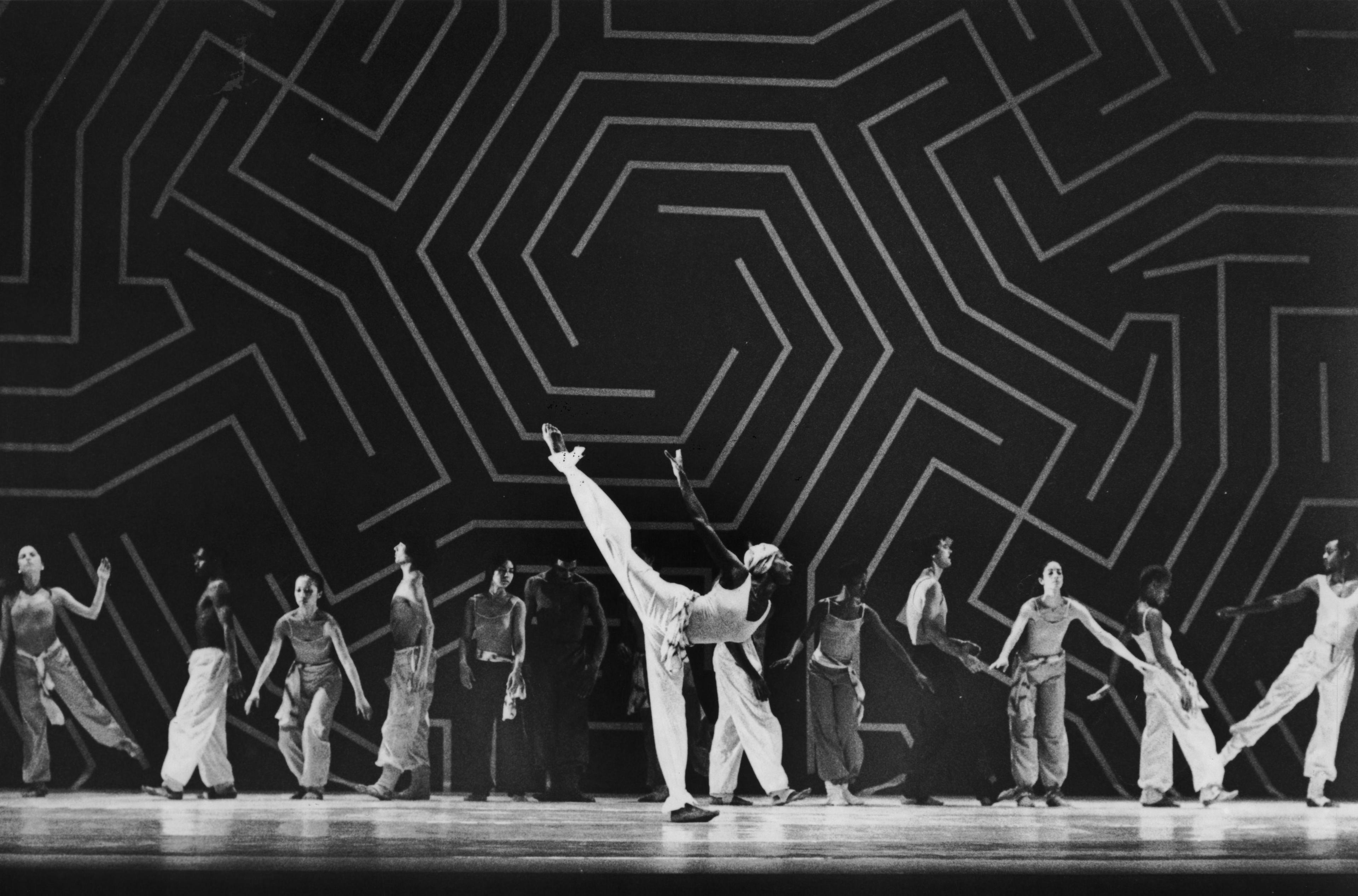  A black-and-white photo of Judith Jamison and the company performing in Caravan, taken by Johan Elbers. The dancers are arranged in a dynamic formation on stage, with one dancer executing a high leg lift, set against a backdrop featuring a bold, geometric pattern.