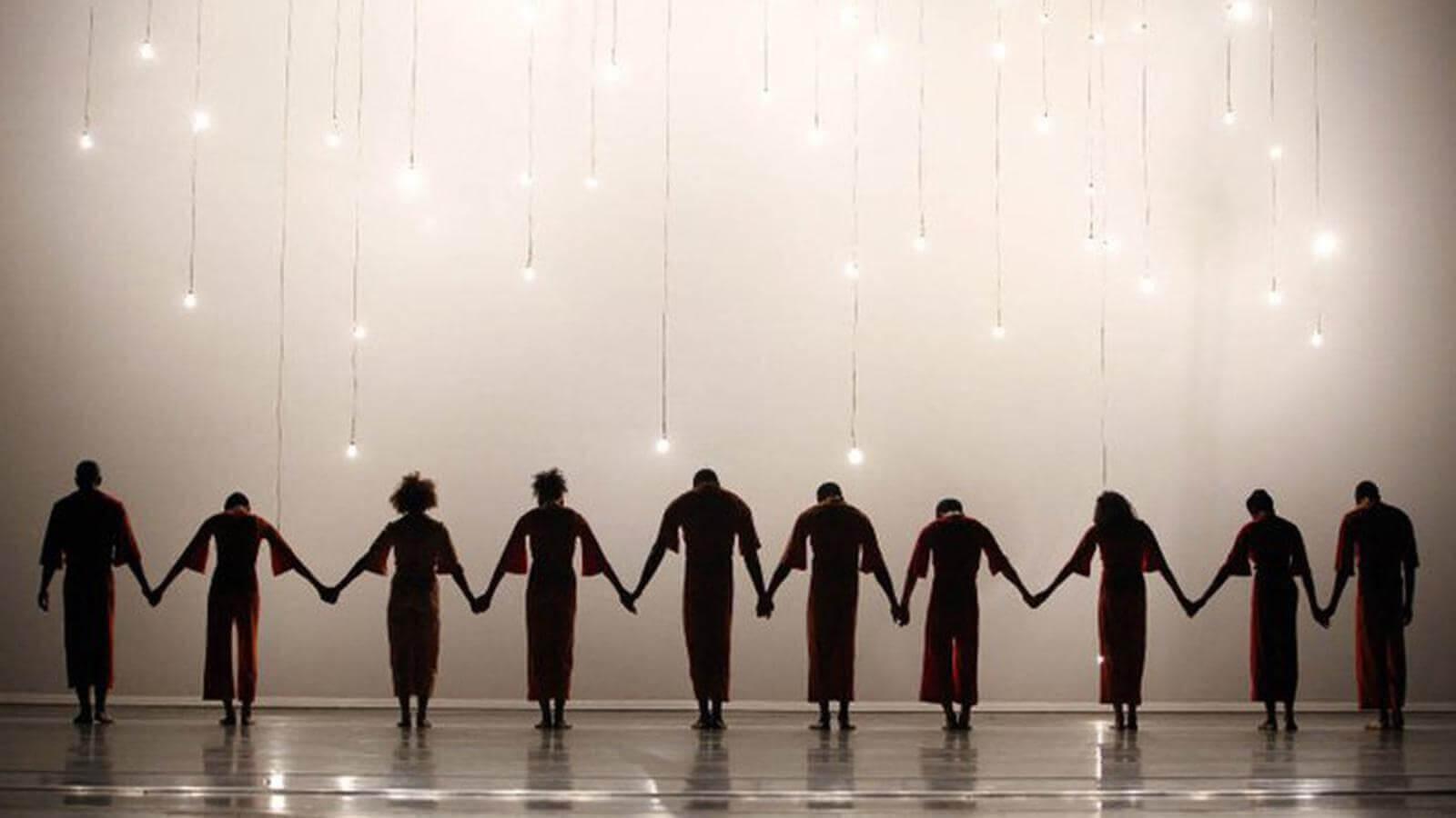 A line of dancers in dark clothing holding hands on stage with hanging light bulbs glowing above them. The background is a soft white.