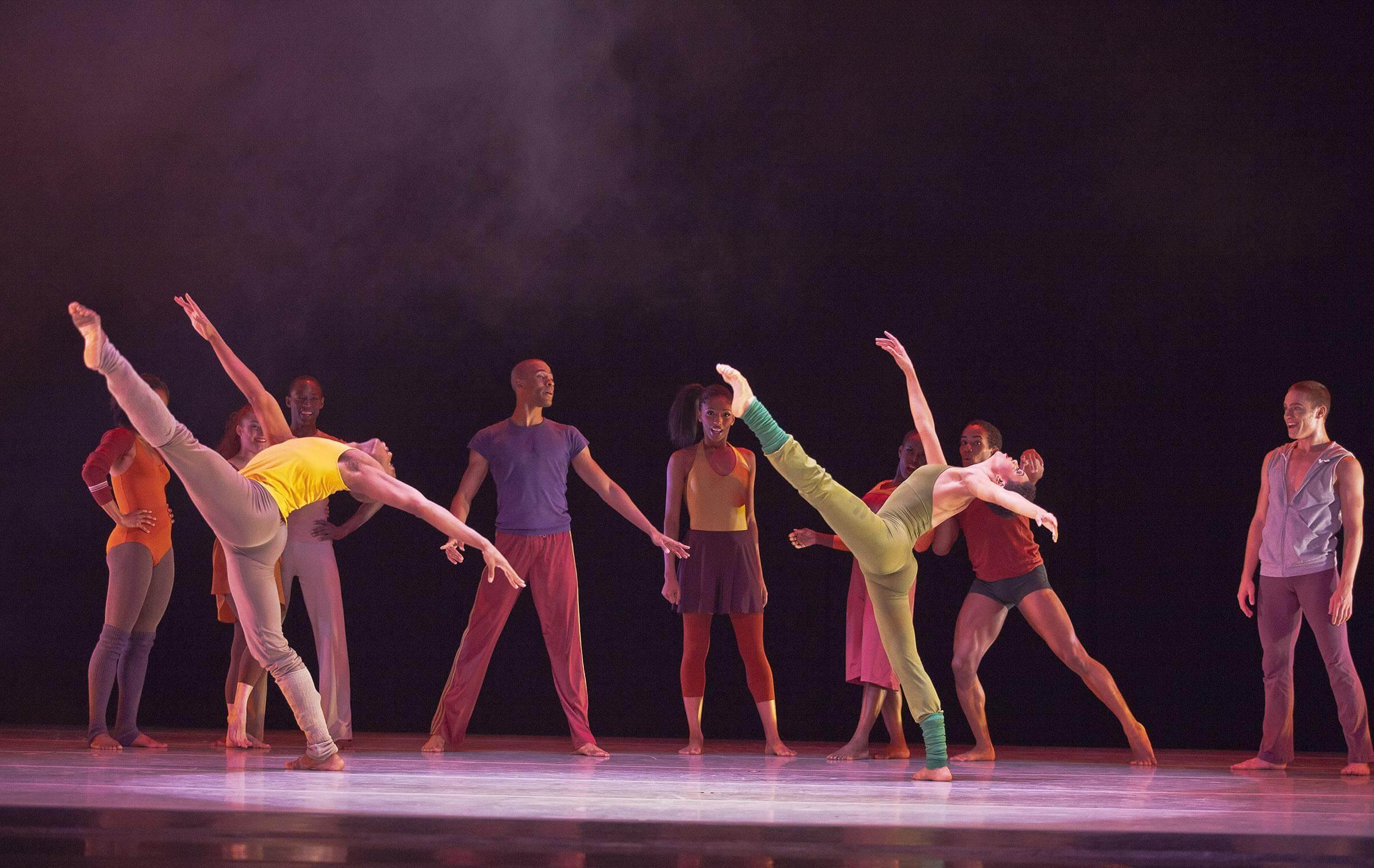  A group of dancers on stage, with two dancers in the foreground performing high leg extensions. They are wearing colorful costumes in shades of yellow, green, orange, and purple. The background is dark.