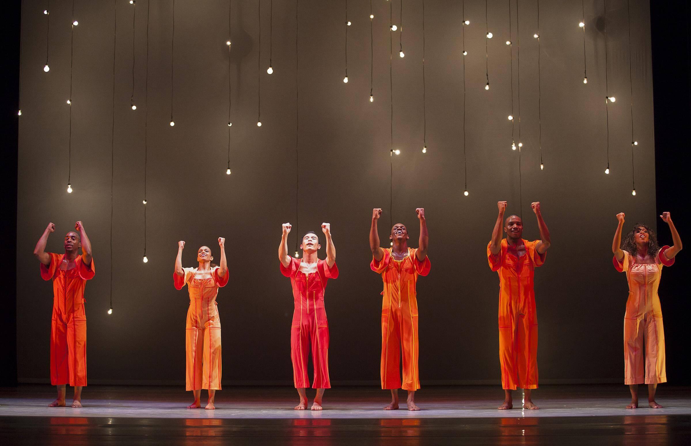 Six dancers in bright orange outfits stand on stage with their arms raised. Above them, numerous light bulbs hang and glow softly against a dark background.