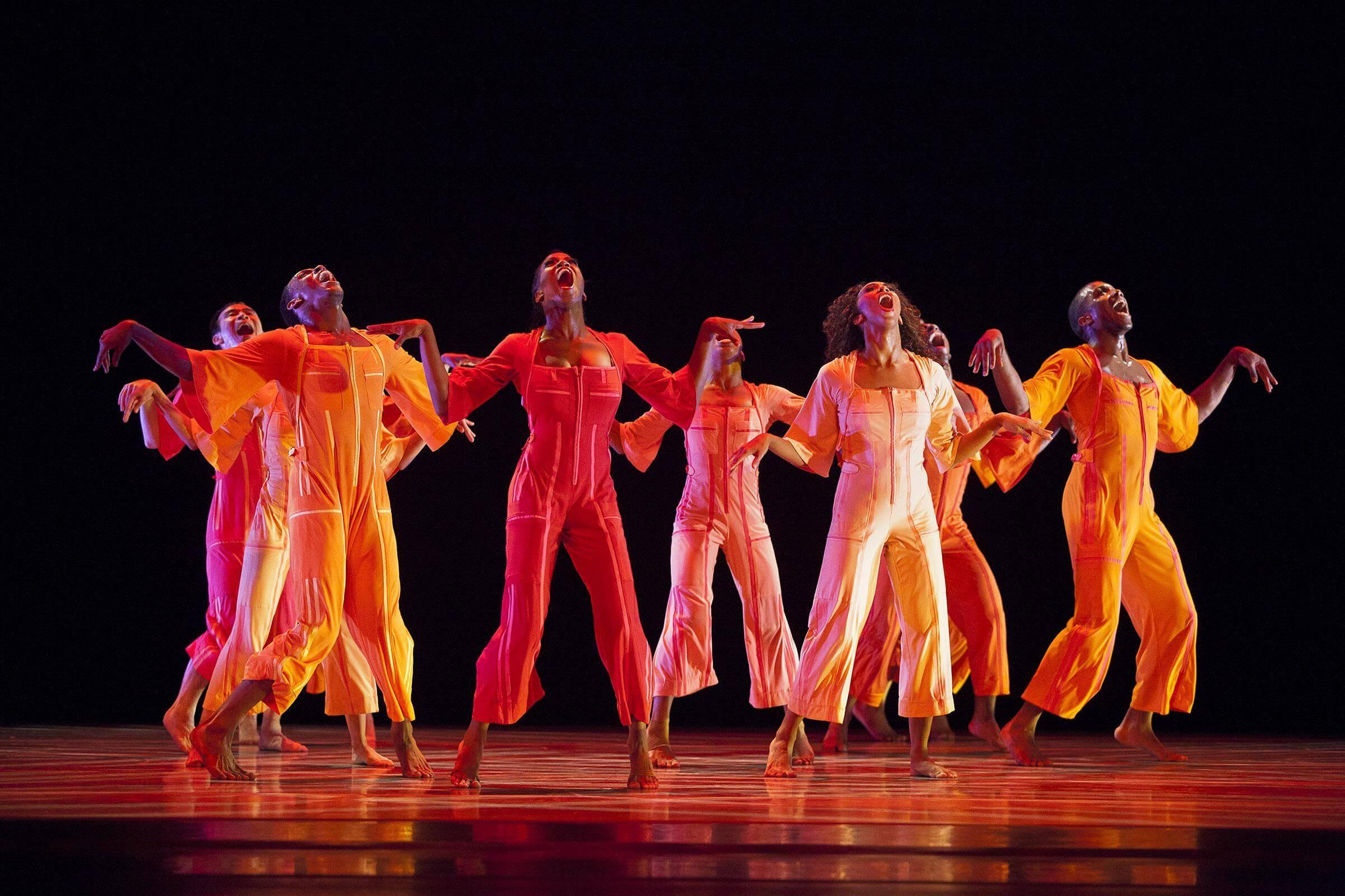  ChatGPT A group of dancers in bright orange and red outfits perform on stage, with their arms raised and mouths open in expressive poses. The background is dark.