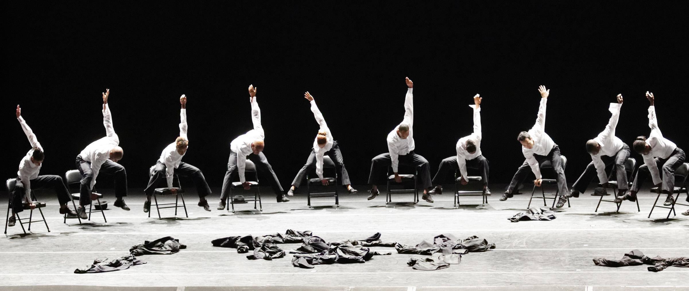 Ten dancers in white shirts and black pants seated on chairs in a line, leaning forward with one arm raised. Discarded black jackets on the floor in front of them.