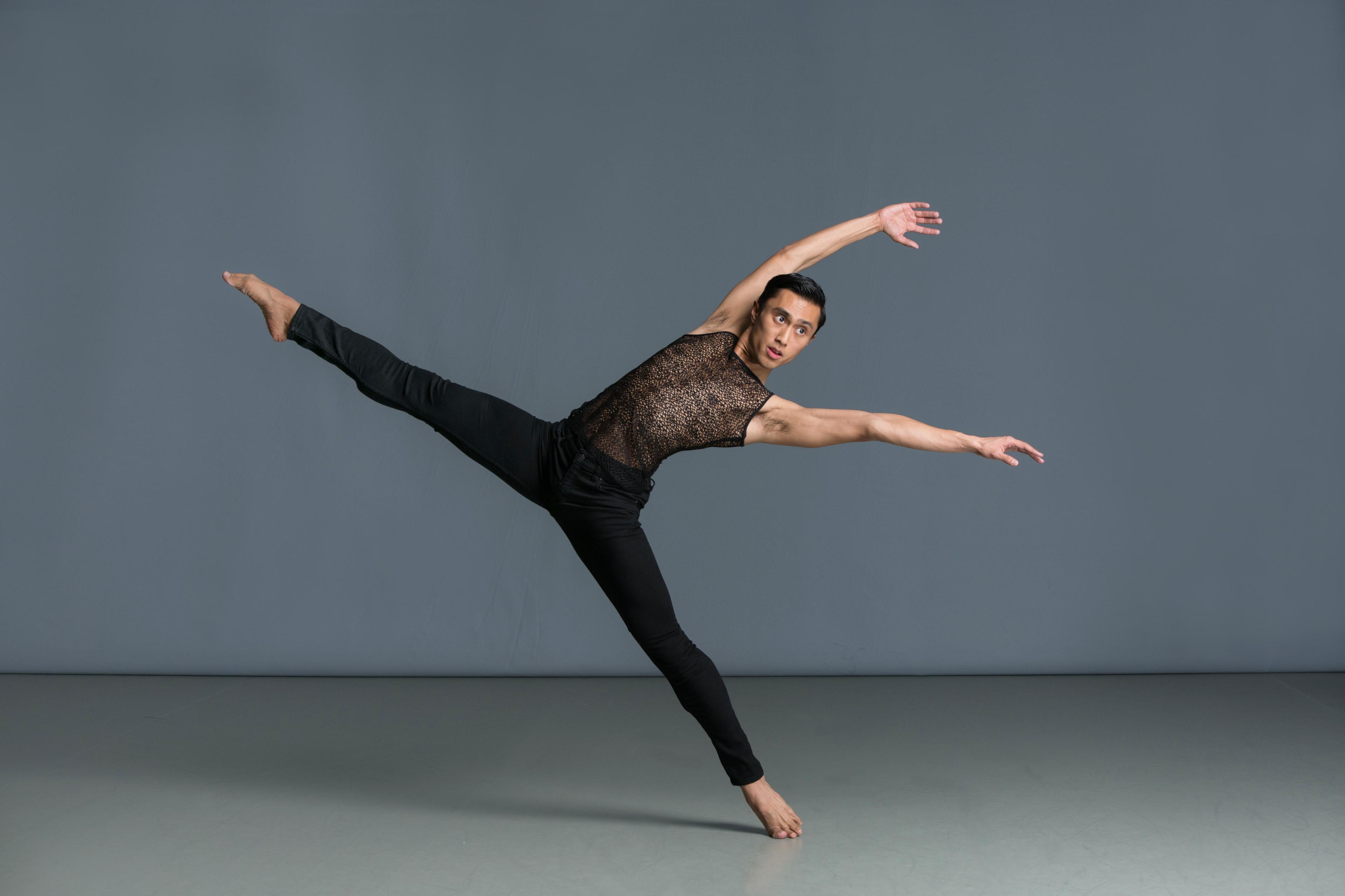 A dance student posing with one leg up and both arms up in black pants and a brown top.