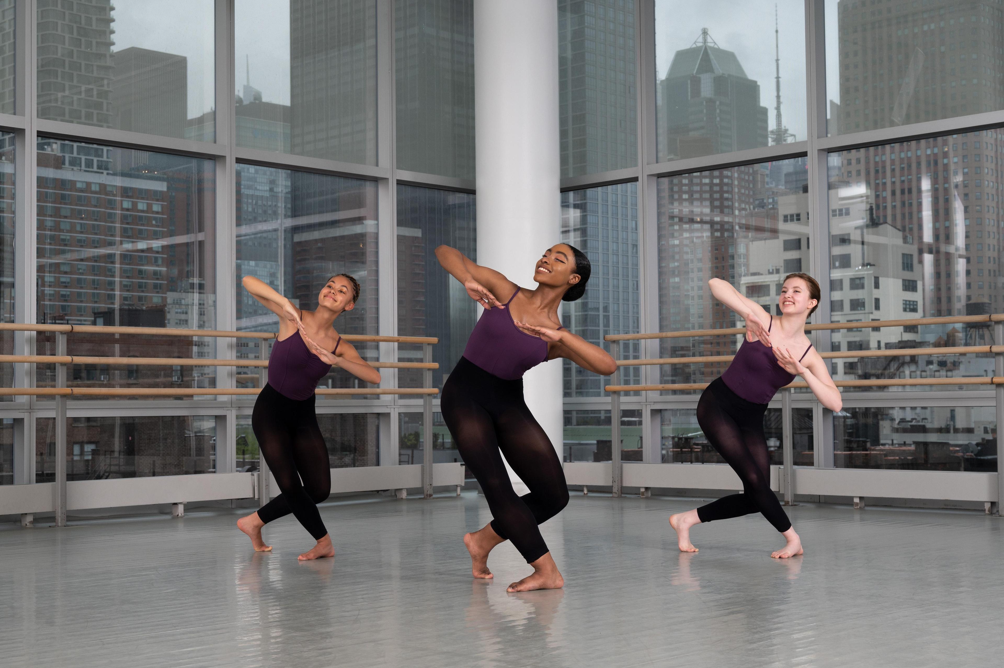 Three girls standing in a triangle on bended knees leaning to their left side and looking up. They are all wearing purple leotards with black tights over them and bare feet.