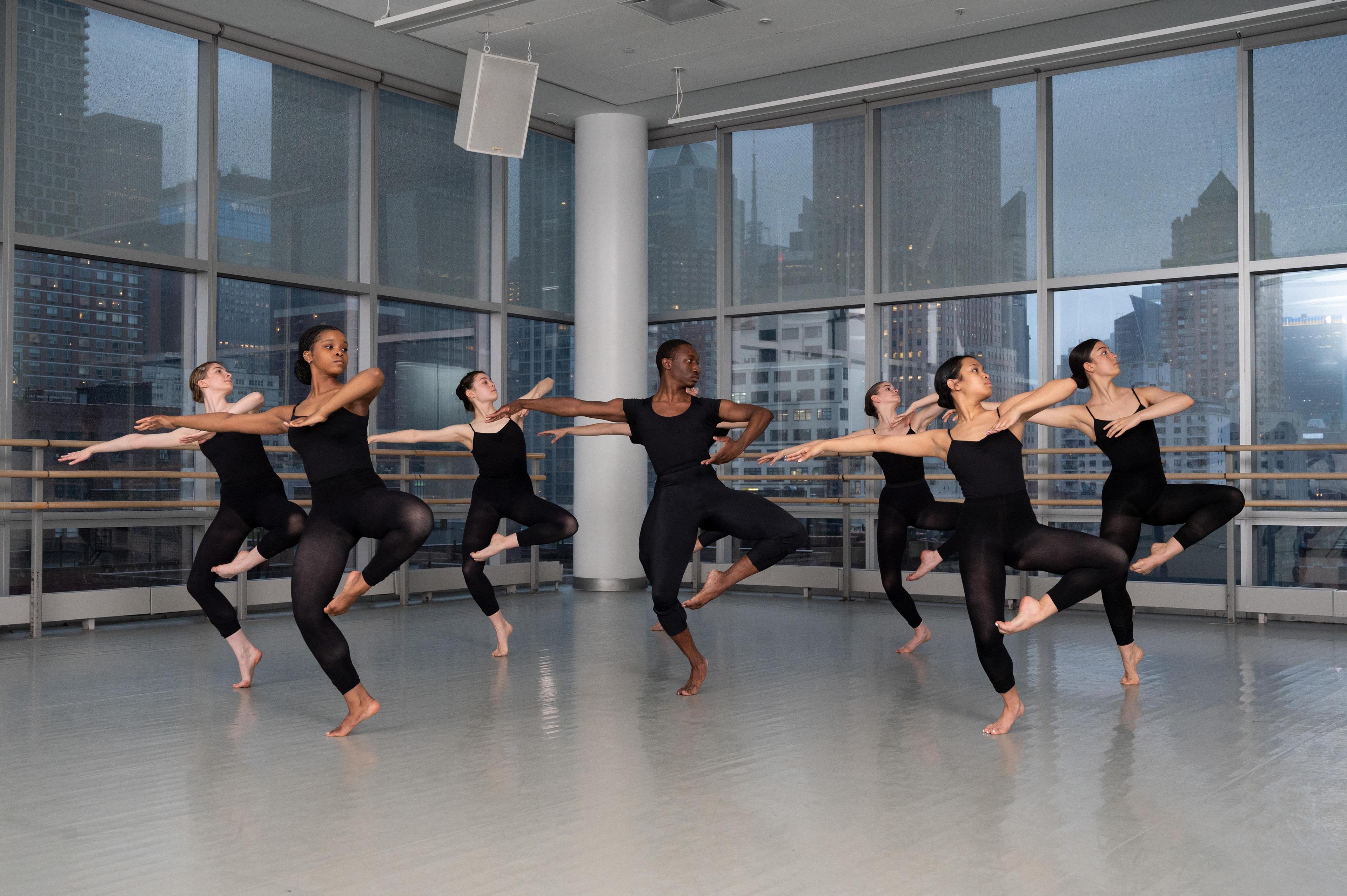 A group of young dance students stand on one leg, wearing black leotards and black tights.