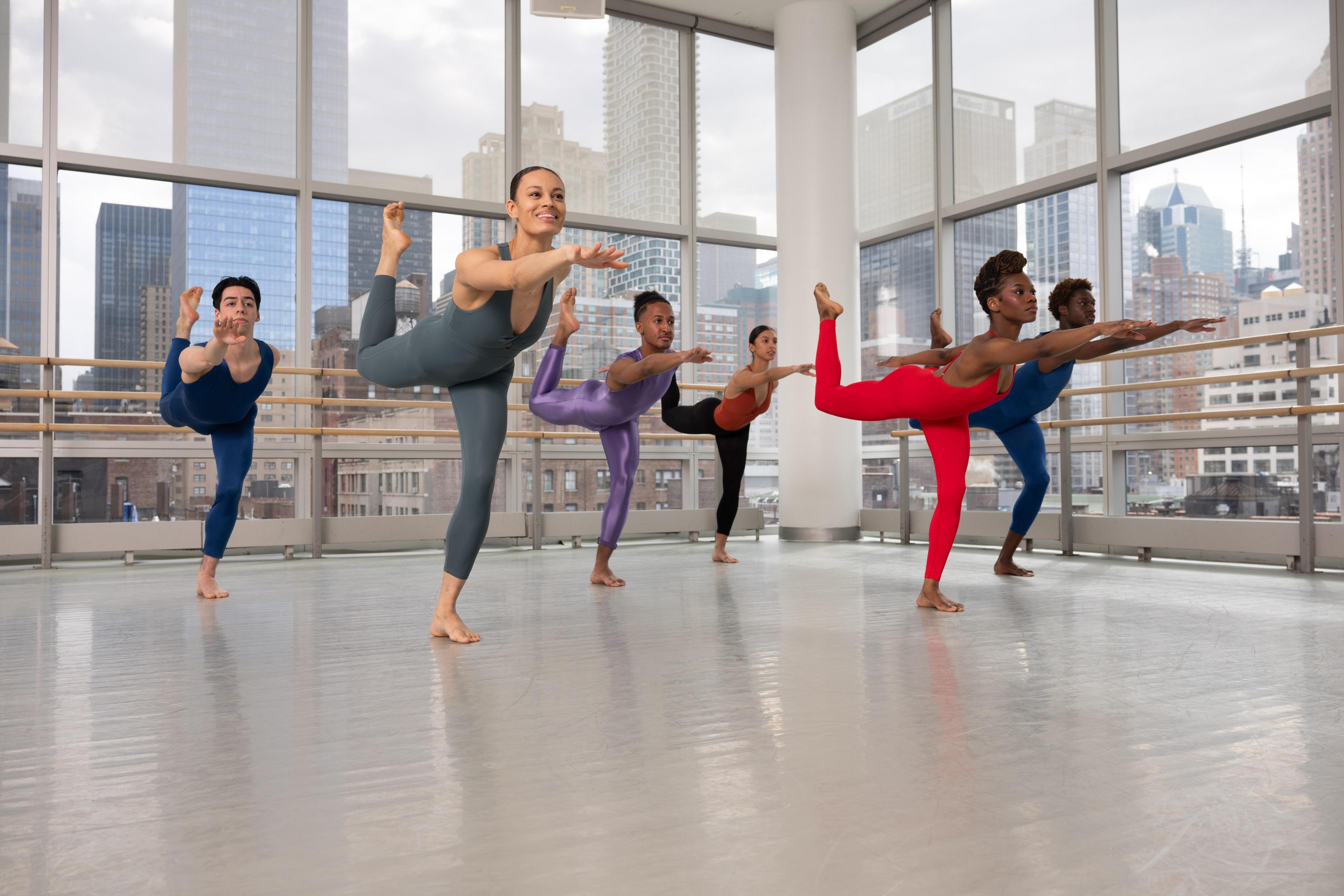 Six dancers in different colored unitards in a dance studio. They have one leg up and are leaning forward with one arm out.