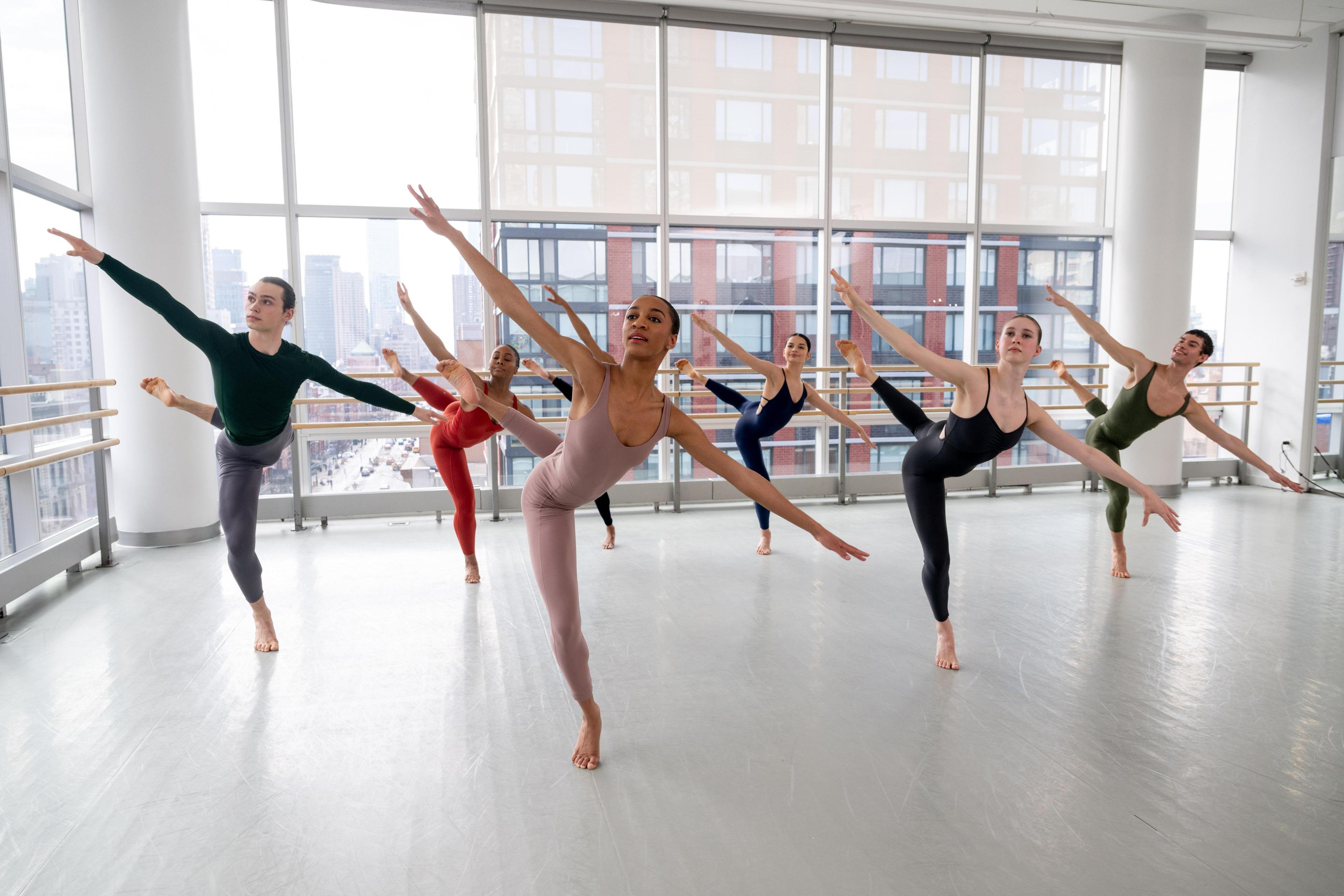 A group of students in various color unitards stand on one leg, with their back leg in attitude and arms reach out on a diagonal line.