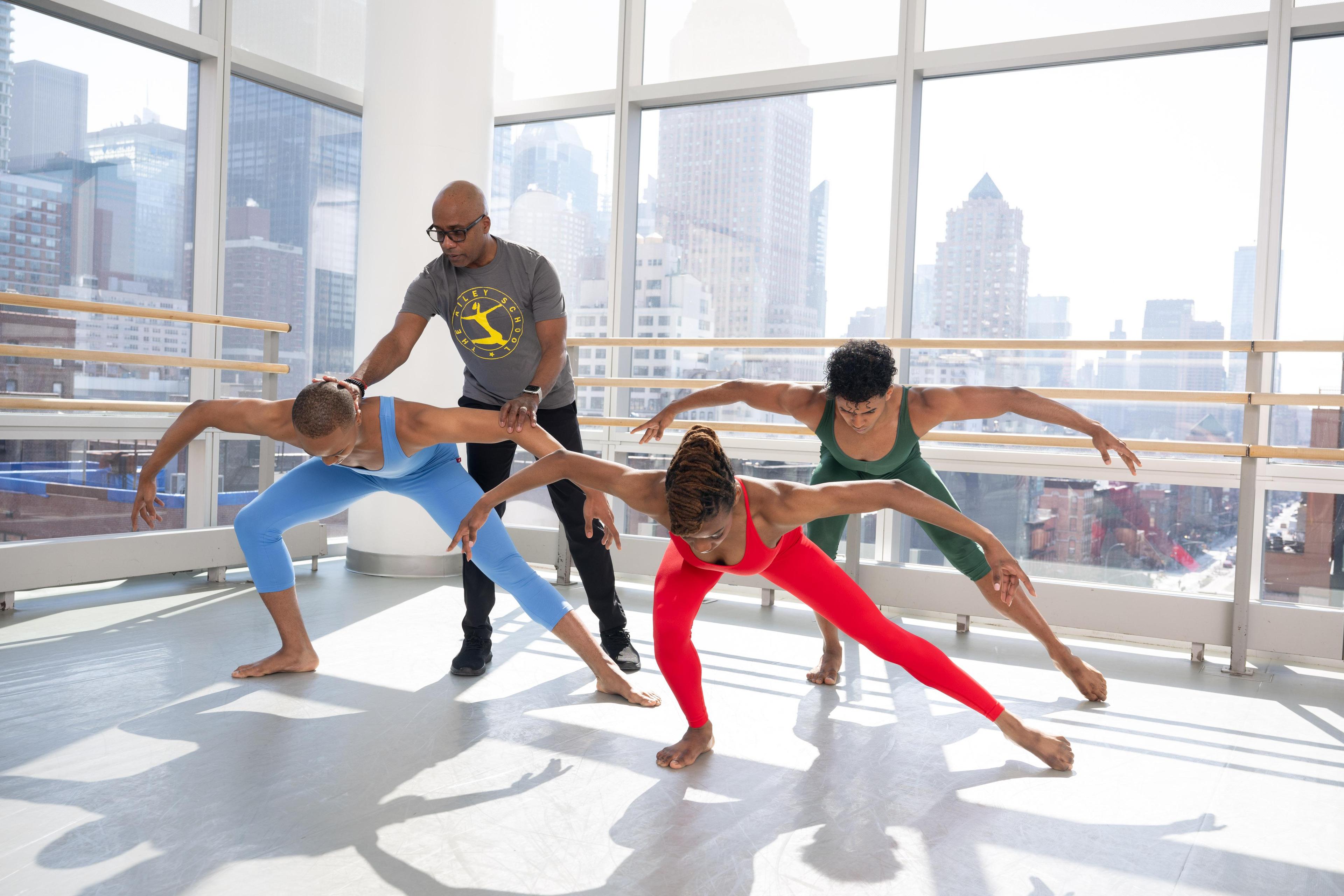 A teacher gives three students a correction on a dance pose wher they have one knee bent, with the other leg extended, looking down at the ground with arms reaching out to their sides making an arch shape.