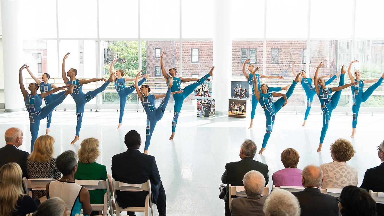 A group of dancers in blue outfits all kick in unison in a dance studio. There is a crowd watching them on chairs.