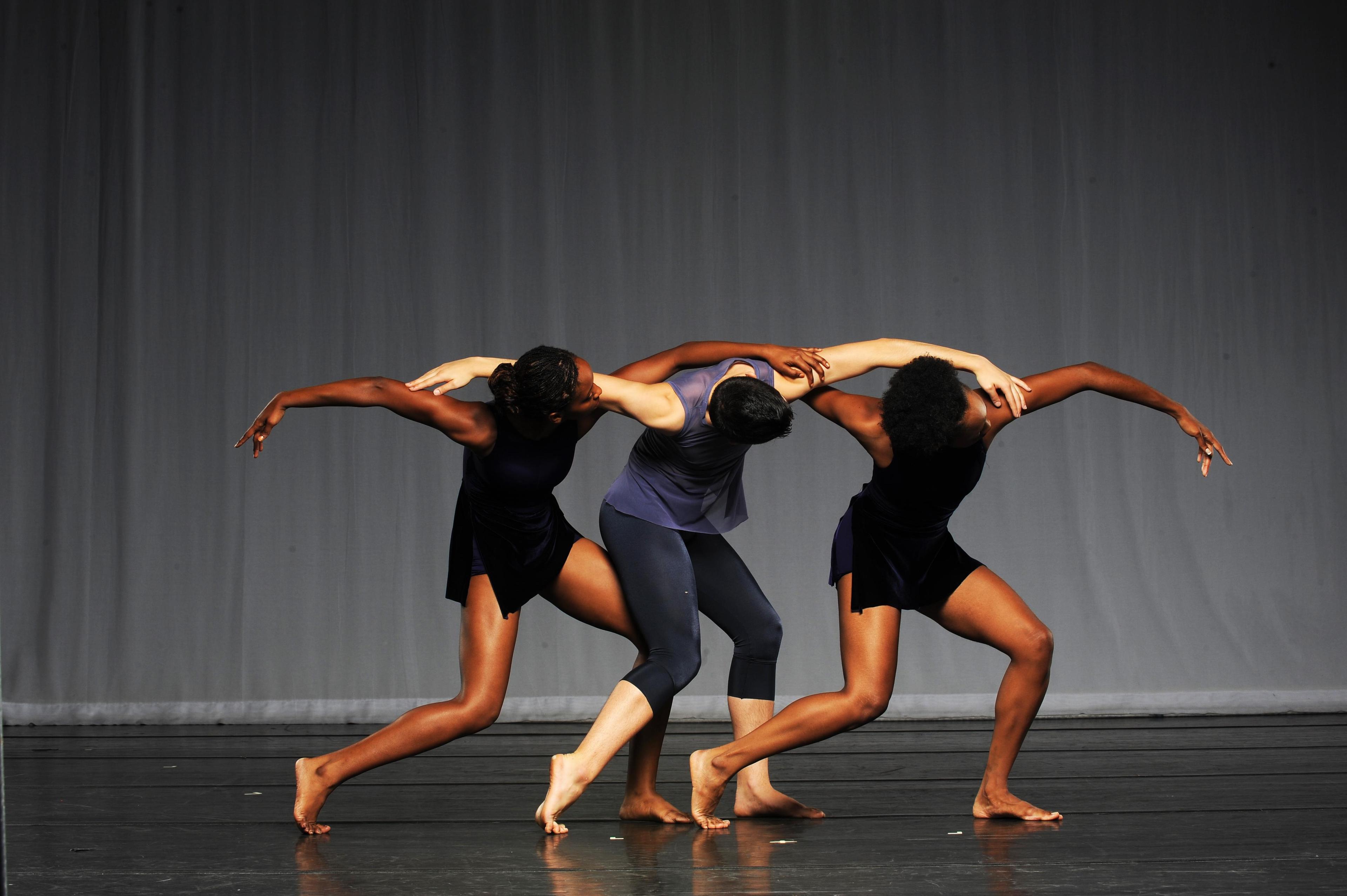 3 students in dark colors with their heads down and arms curved around one another and one foot popped