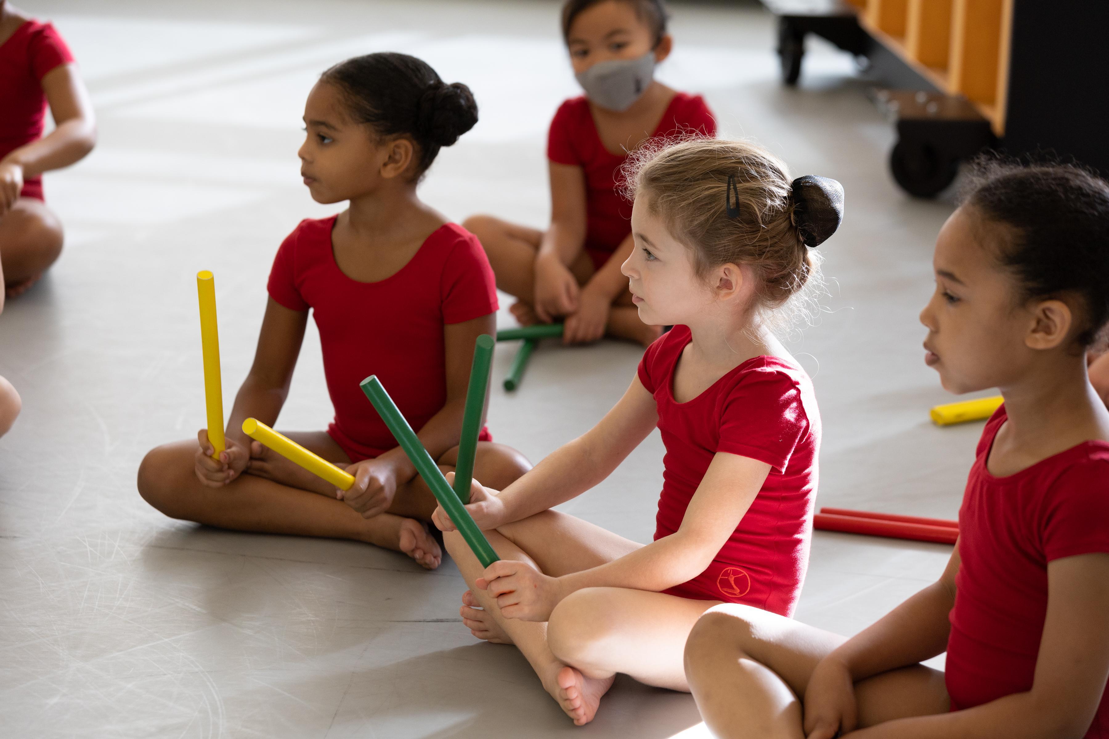 A group of girls in red leotards sit cross legged on the floor.