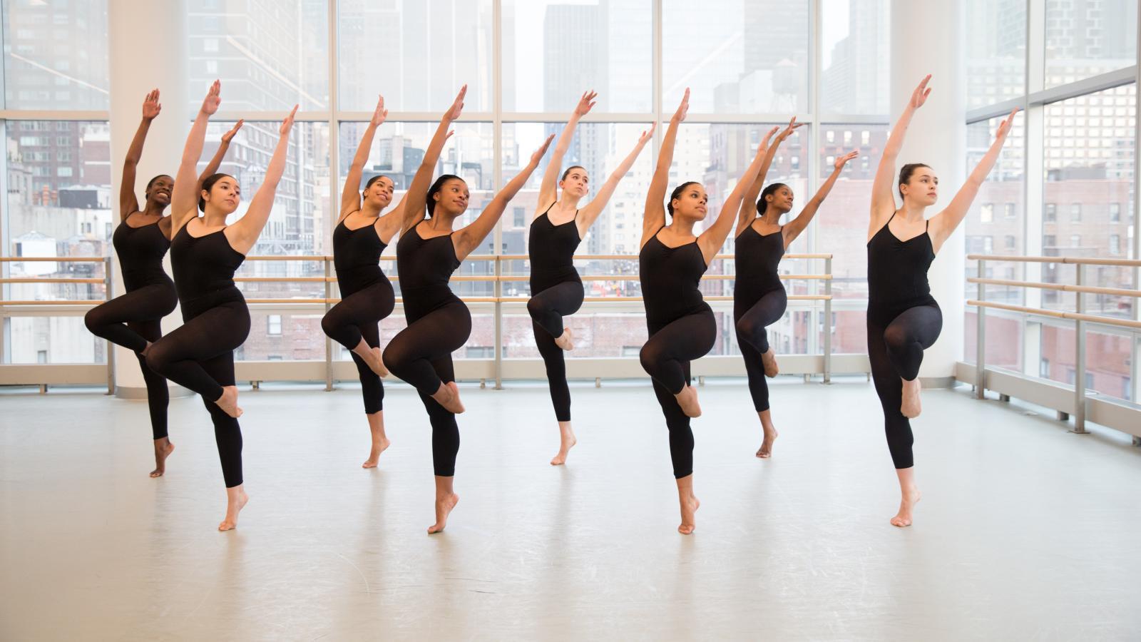 Dancers in black leotards are in a studio. They are all in sync with one knee up and their arms raised