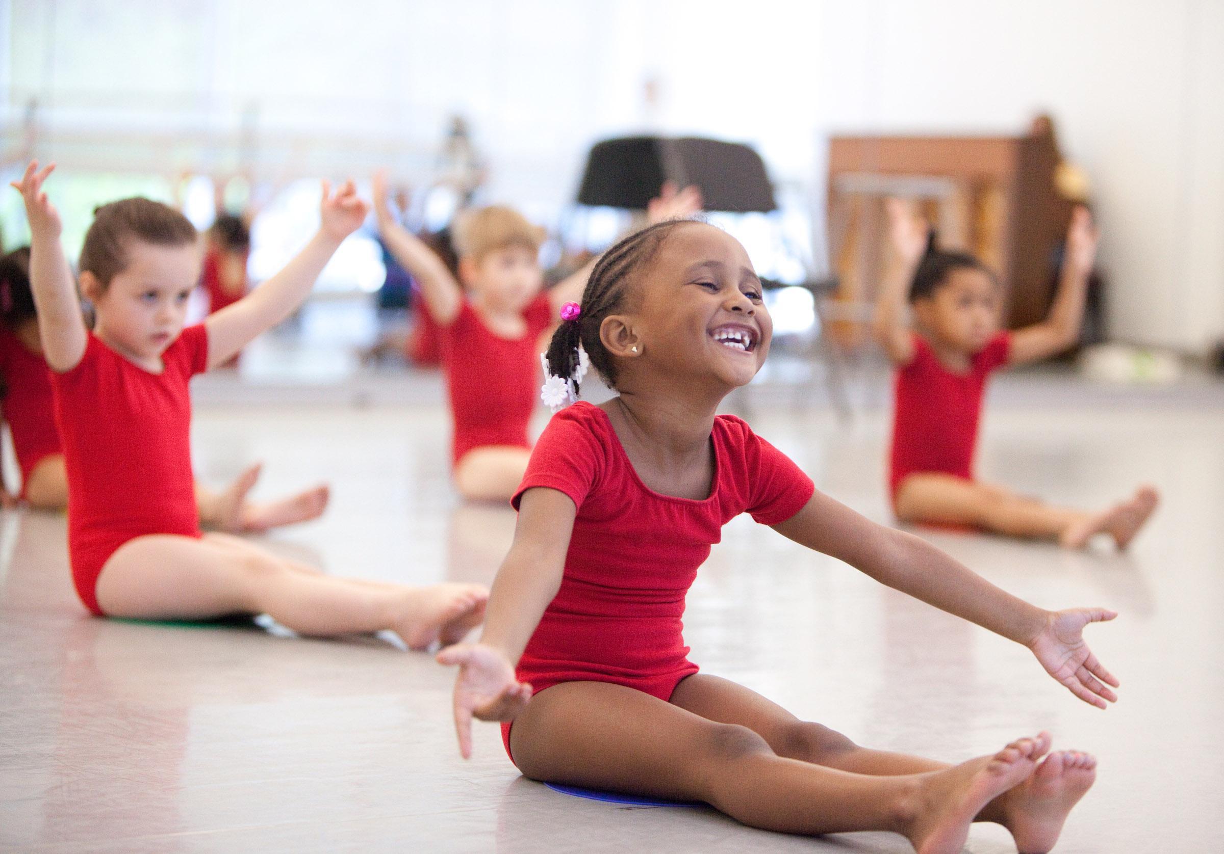 Little girls in red leotards sit on the floor with arms raised in a studio