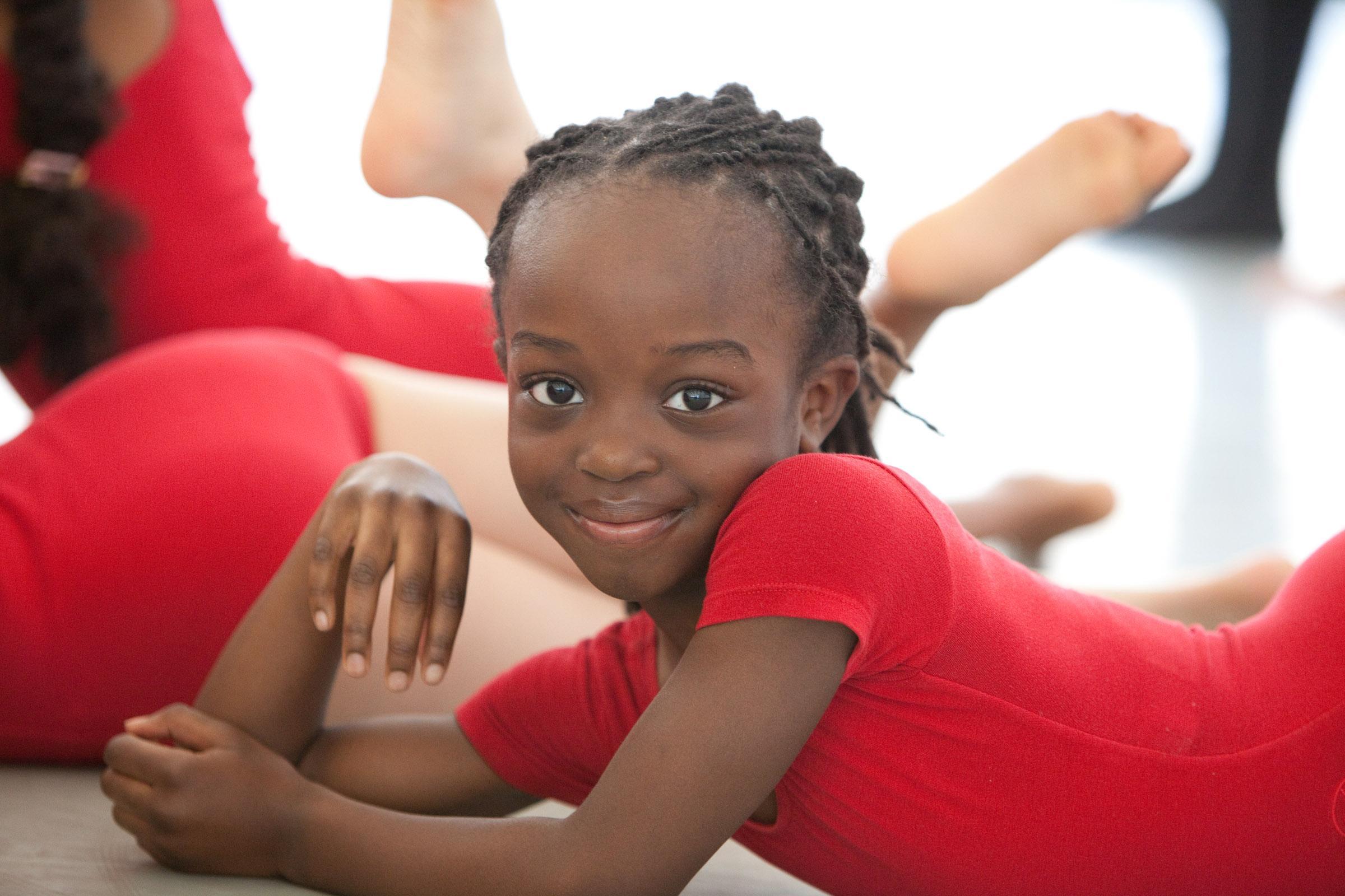 A little girl with cornrows in a red leotard poses on the ground