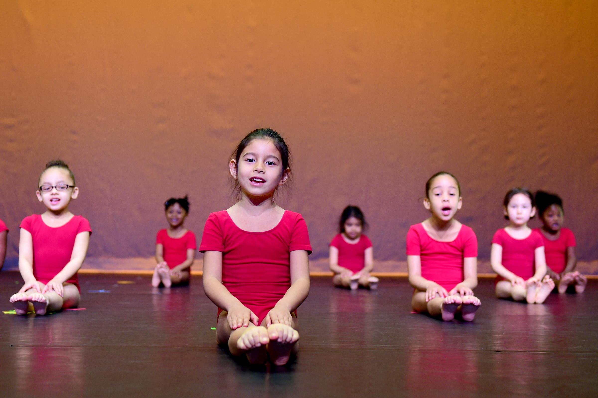 Little girls in red leotards sit on stage with their legs in front of them