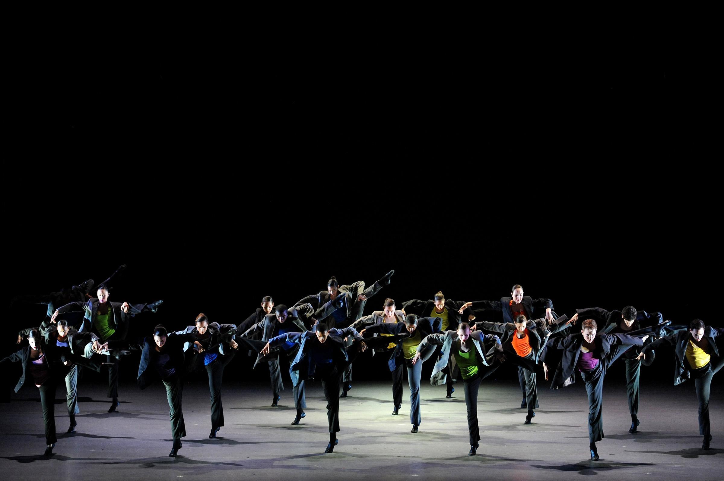 A group of dancers in black suits posing on stage standing on one leg