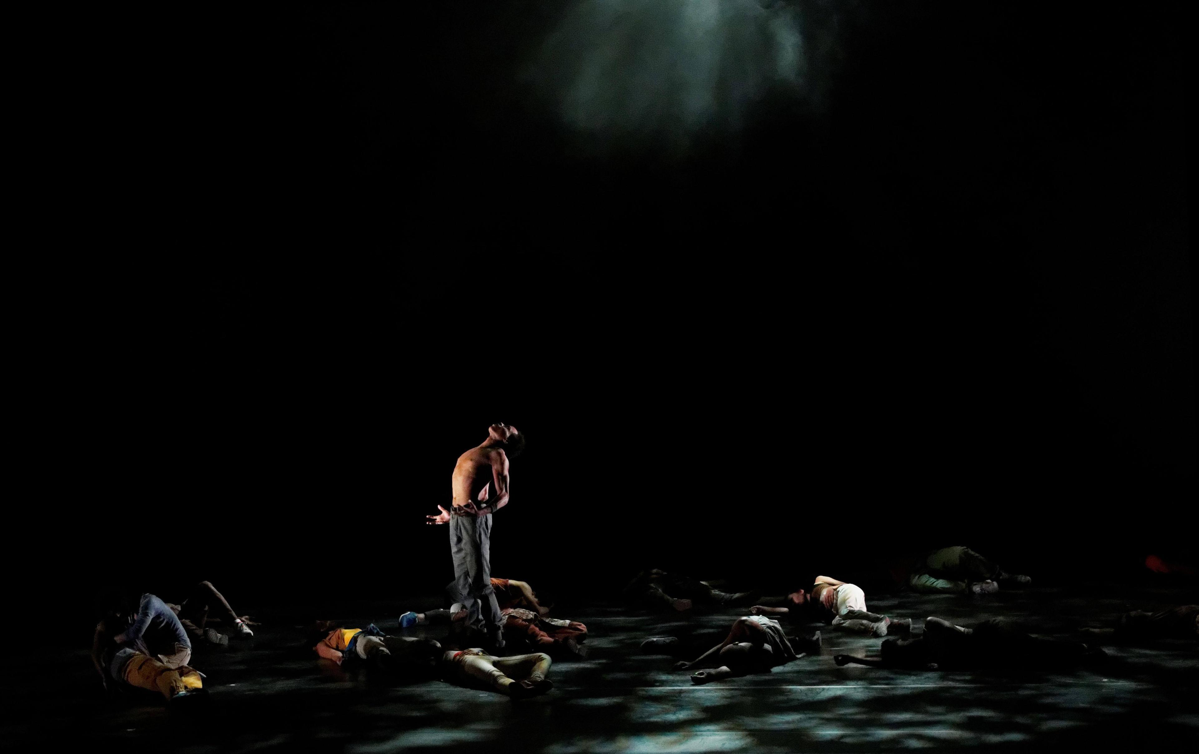 A dimly lit stage with a spotlight highlighting a shirtless dancer standing and arching backward. Several other dancers lie on the floor around him, partially illuminated by scattered light.