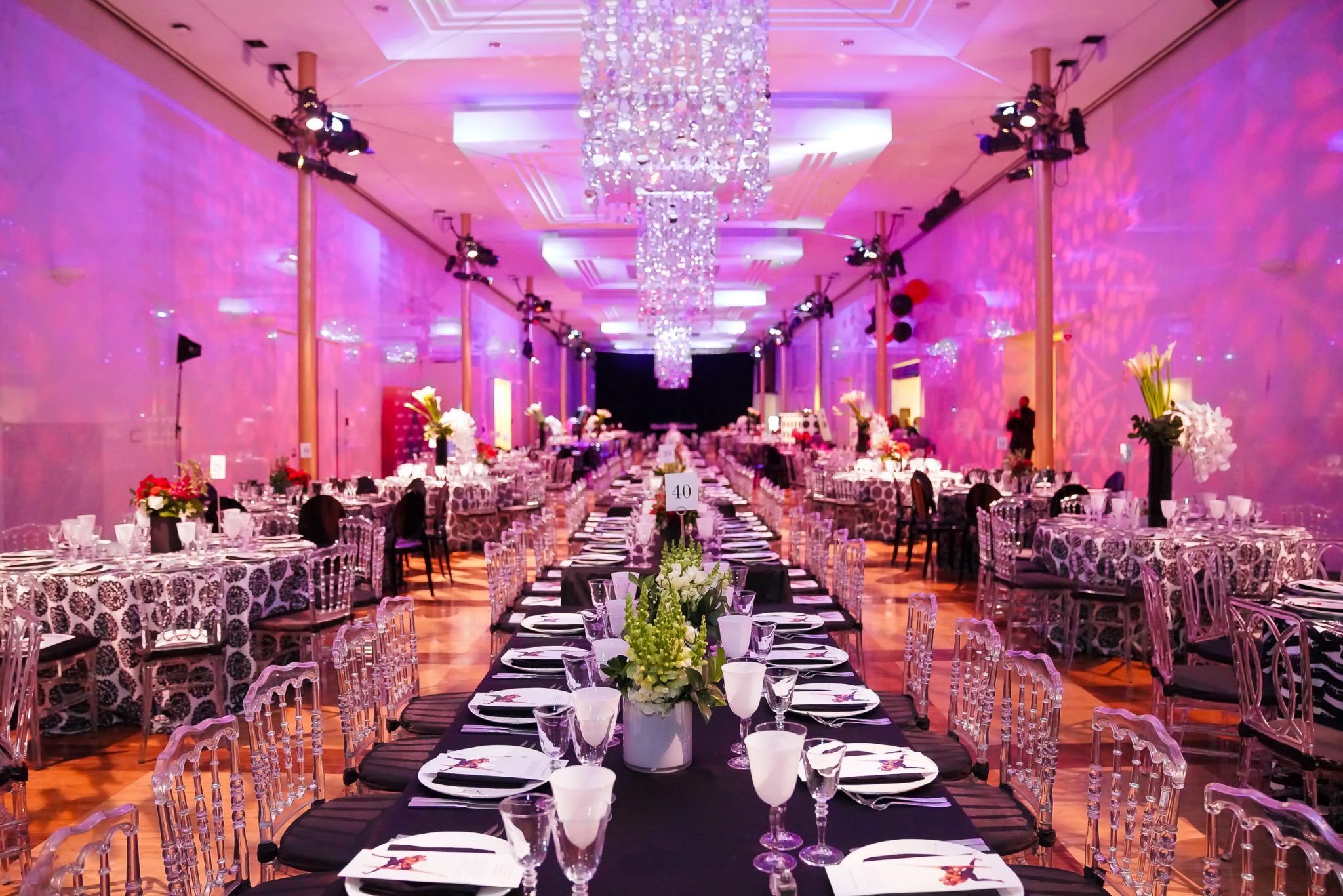 Ballroom with multiple tables with settings and flowers. The room has a pink color due to lighting. There are crystal chandeliers hanging from the ceiling.