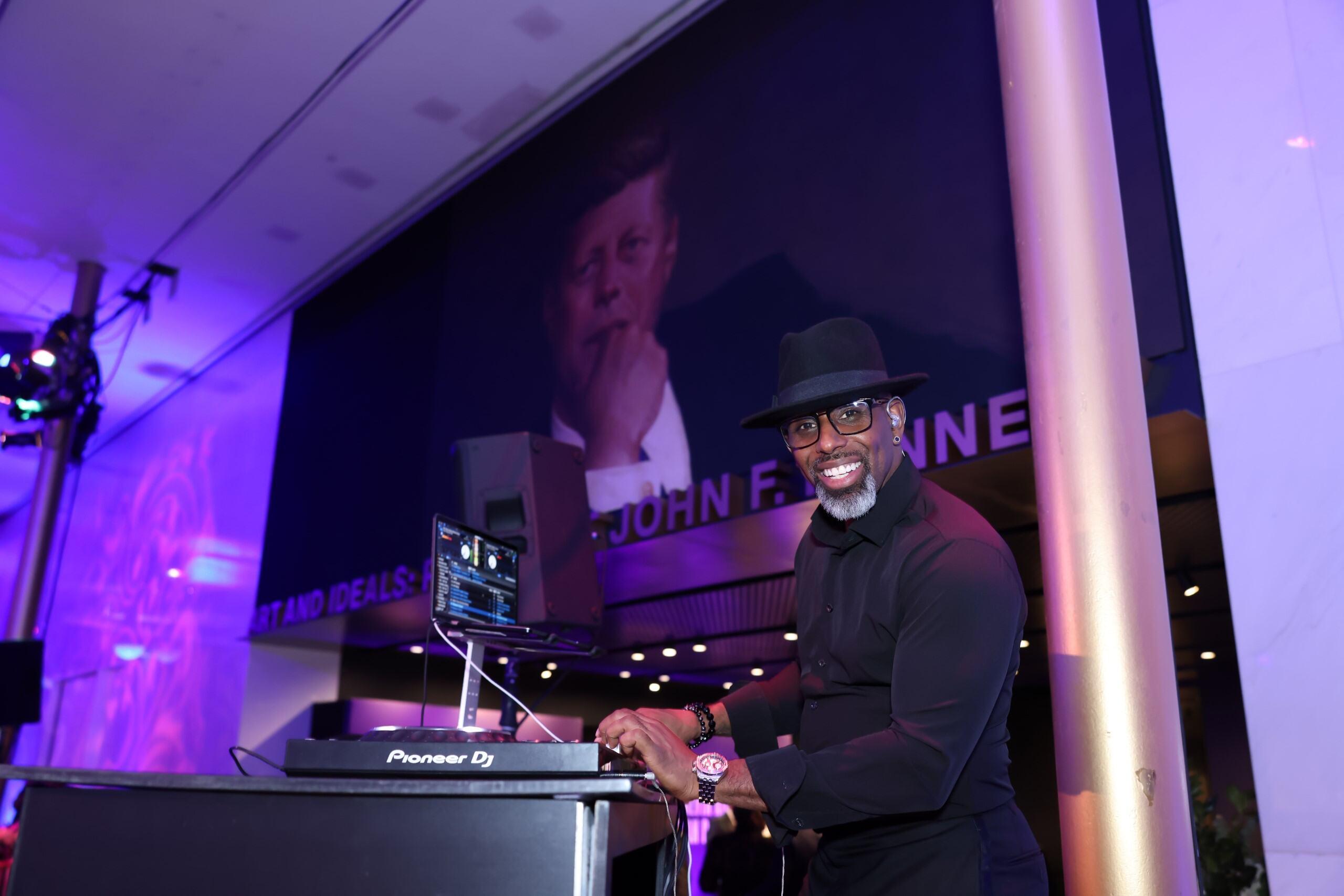 A man in a DJ booth. He is smiling at the camera. The room has a purple tone due to party lights.