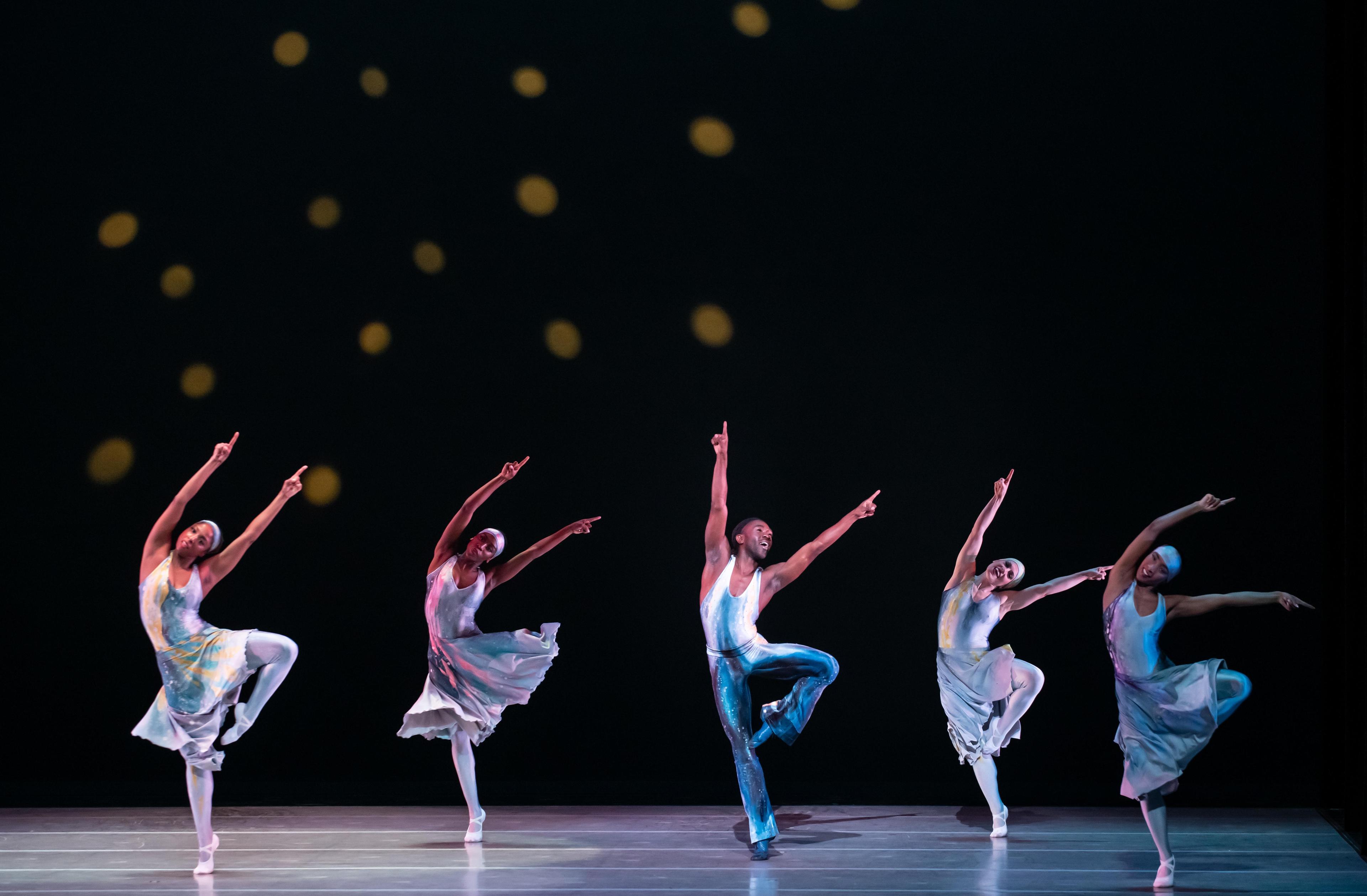 A group of dancers in tie dye costumes stand on one leg reaching arms up into the air.