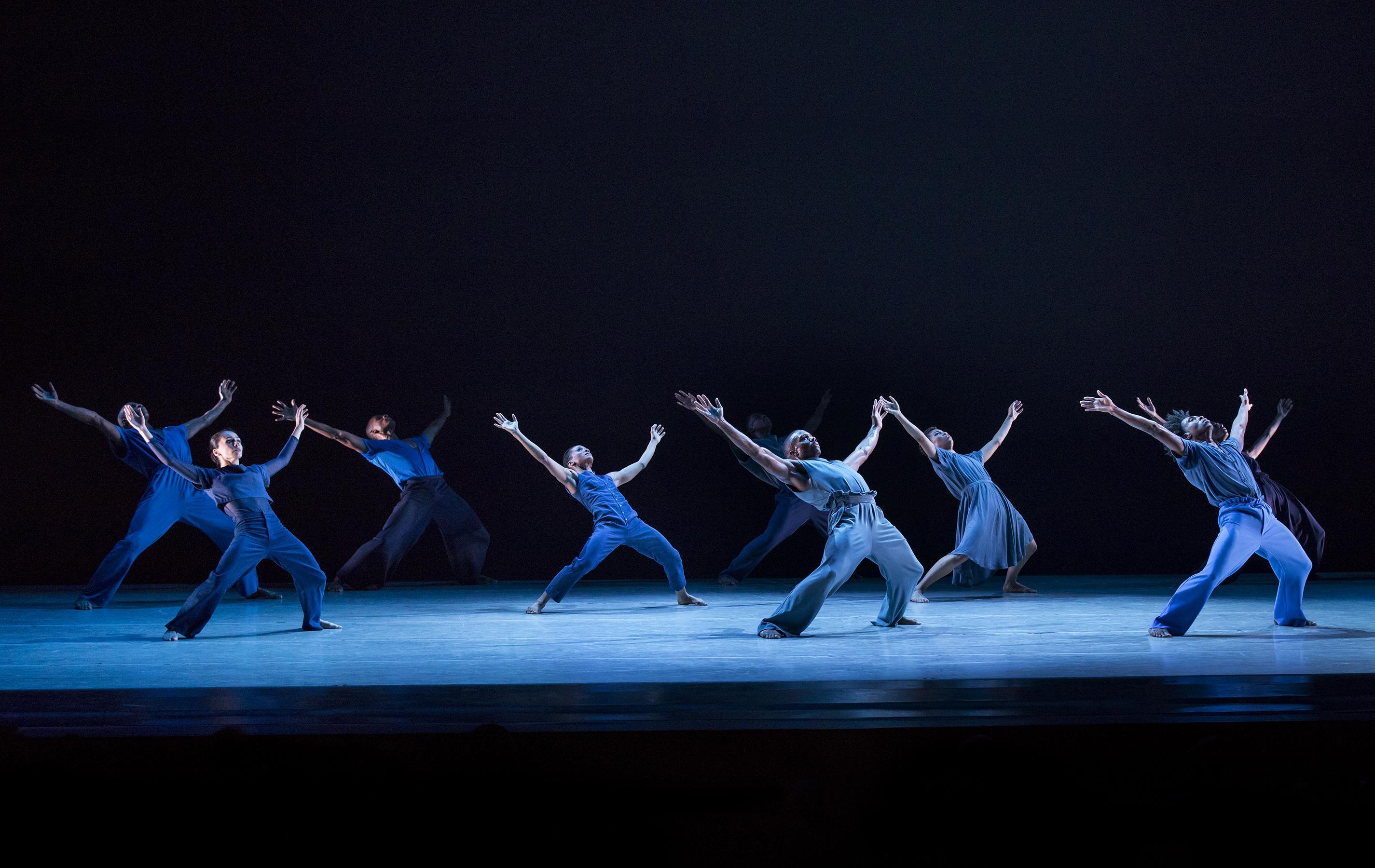 A group of dancers dressed in various shades of blue are scattered around a stage, all leaning backwards with their legs bent