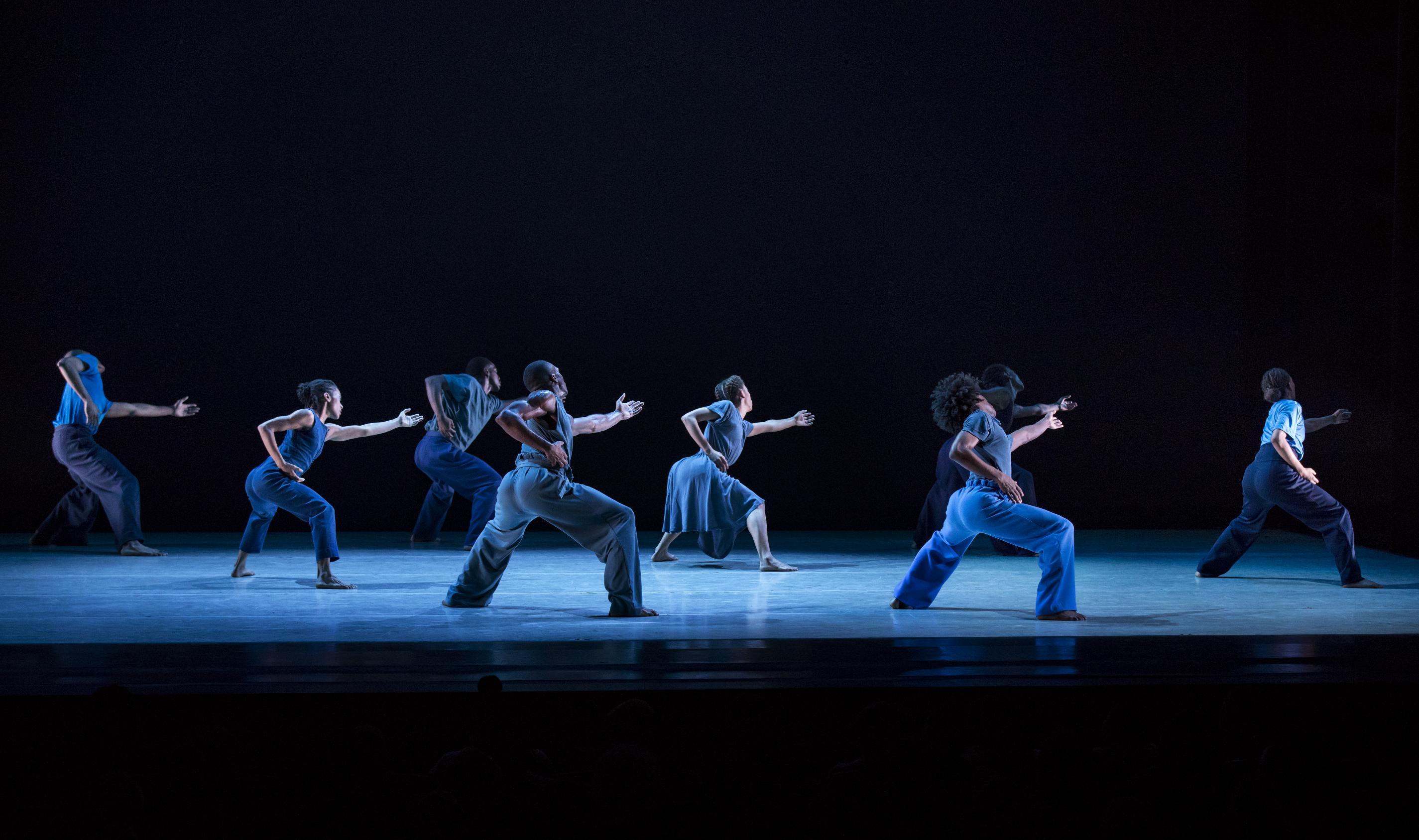 a group of dancers dressed in various shades of blue stand with knees bent and their backs to the audience.