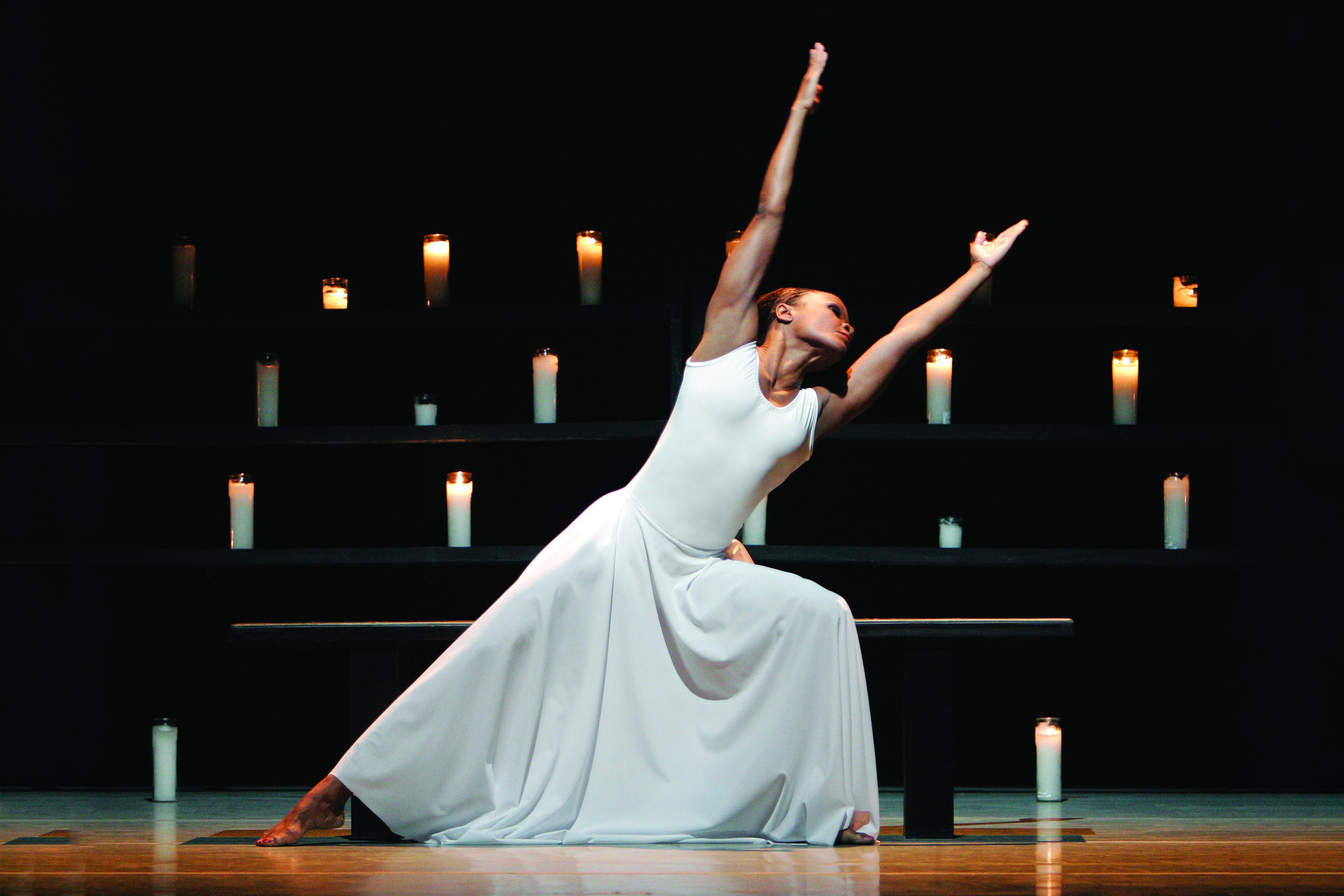 A woman in a white dress is lunging to one side with her arms outstretched. There are rows of candles behind her.