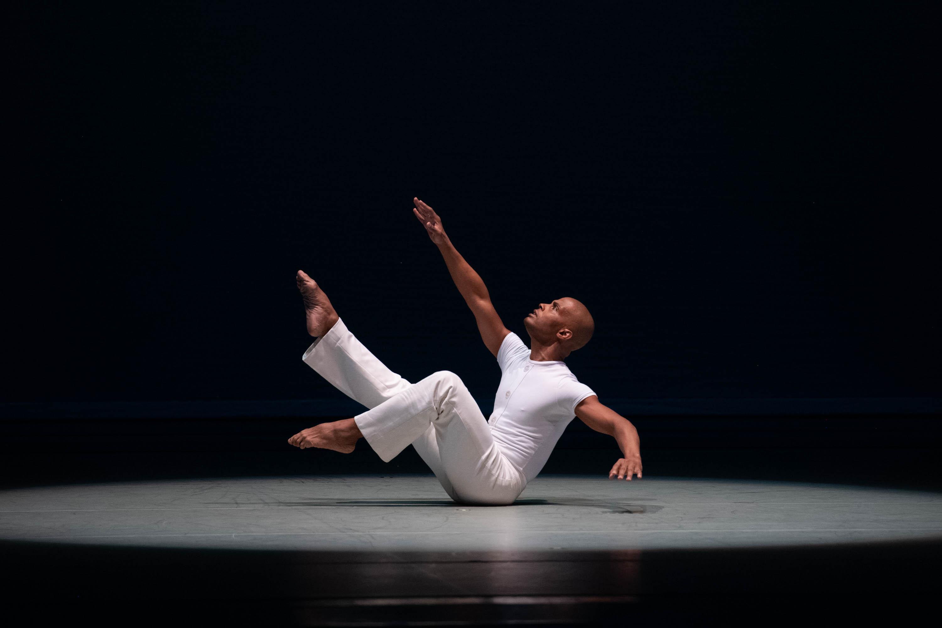 A man in white sits on the floor with his arms and legs reaching up to the sky.